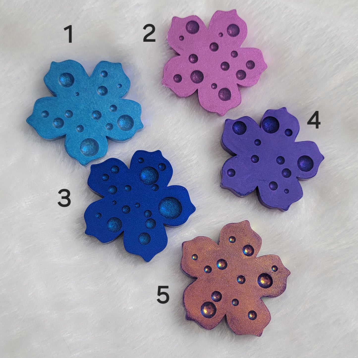 Five flower blossom shaped epoxy resin pieces with dewdrop effect and shimmering metallic finishes in shades of purple and blue.