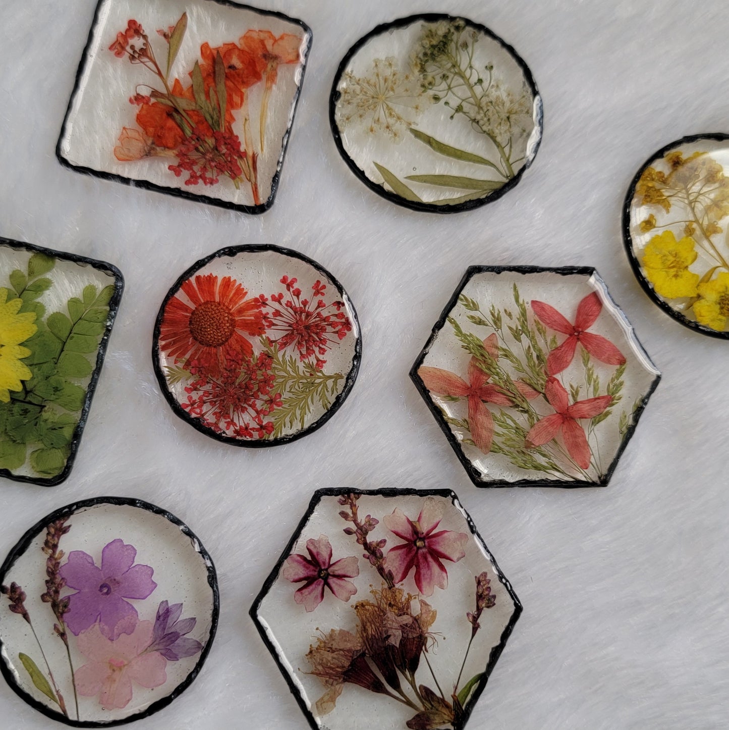 Epoxy resin pieces in varied shapes - square, hexagon, standard round and larger round - each with unique array of pressed flowers and foliage and black pigmented edges.