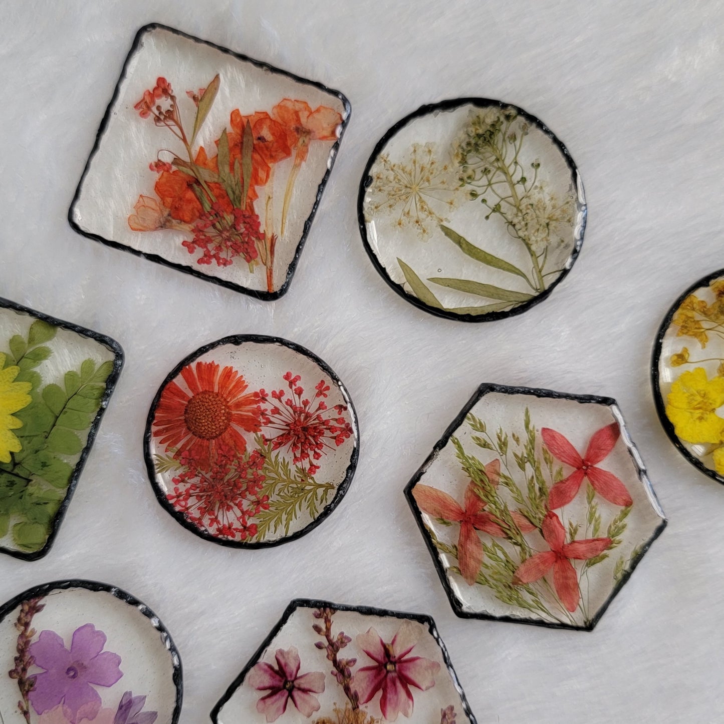 Epoxy resin pieces in varied shapes - square, hexagon, standard round and larger round - each with unique array of pressed flowers and foliage and black pigmented edges.
