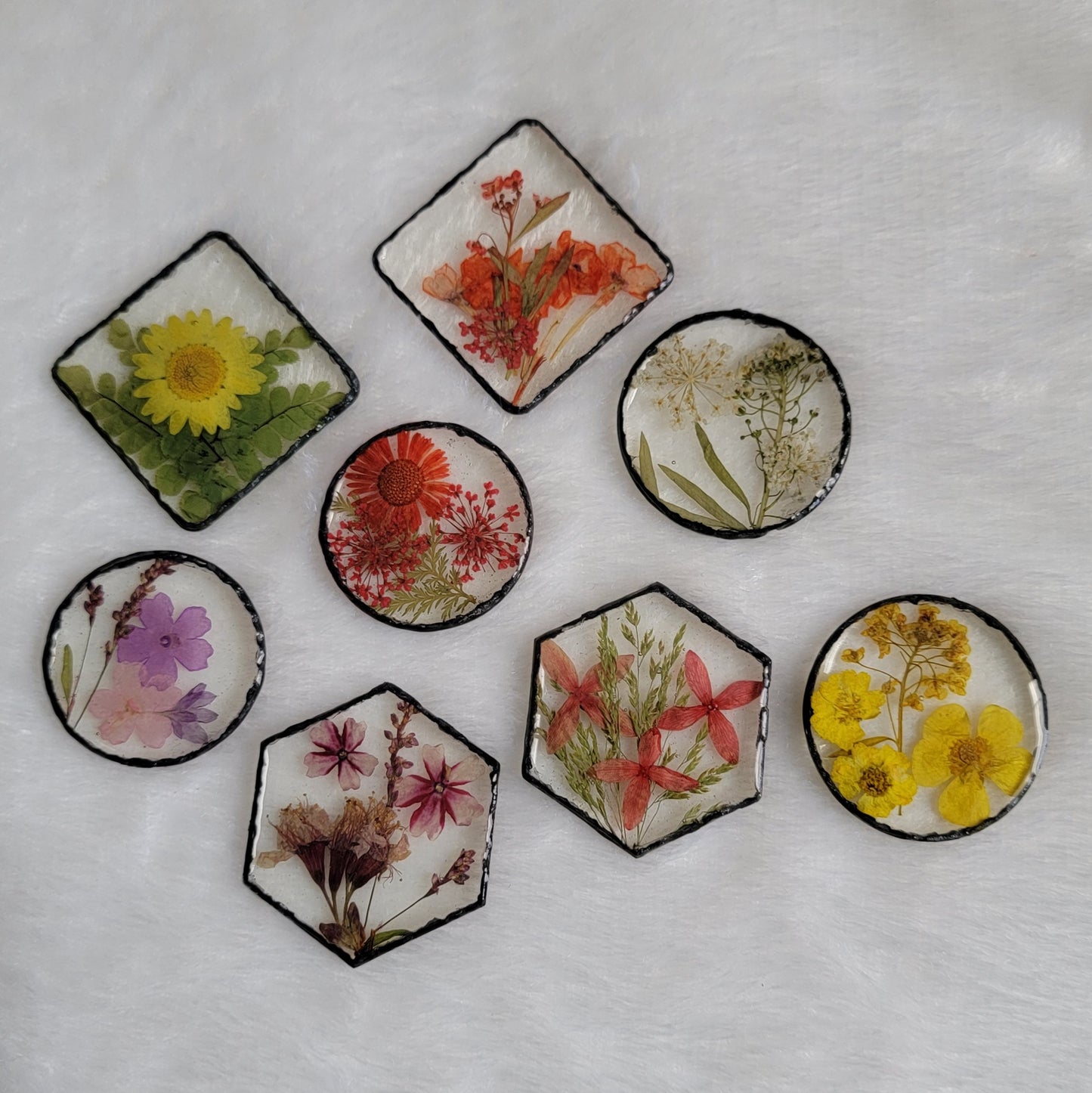 Eight epoxy resin pieces in varied shapes - square, hexagon, standard round and larger round - each with unique array of pressed flowers and foliage and black pigmented edges.