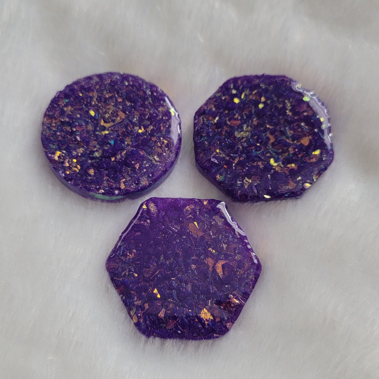 Three epoxy resin pieces - round, octagon, and hexagon - with shimmering golden iridescent crystalline texture encased within and deep purple pigment.