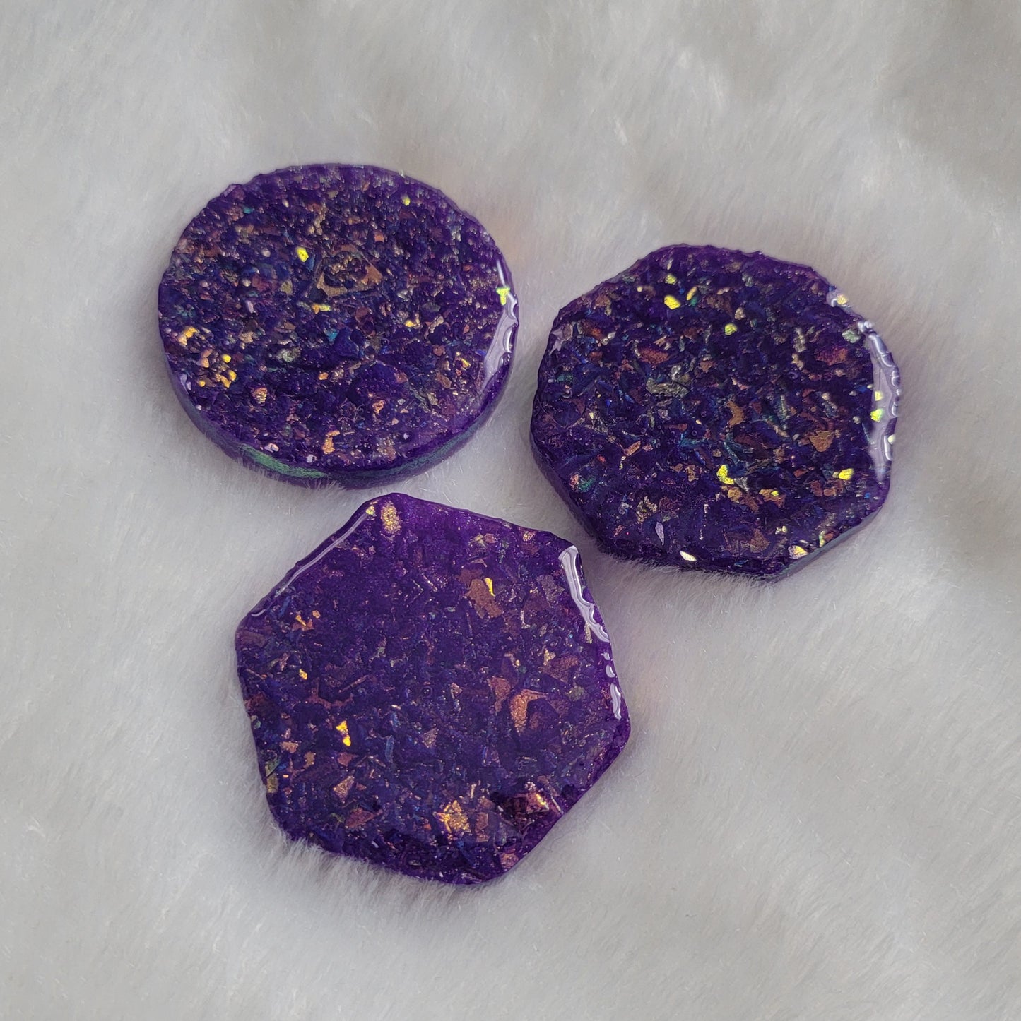 Three epoxy resin pieces - round, octagon, and hexagon - with shimmering golden iridescent crystalline texture encased within and deep purple pigment.
