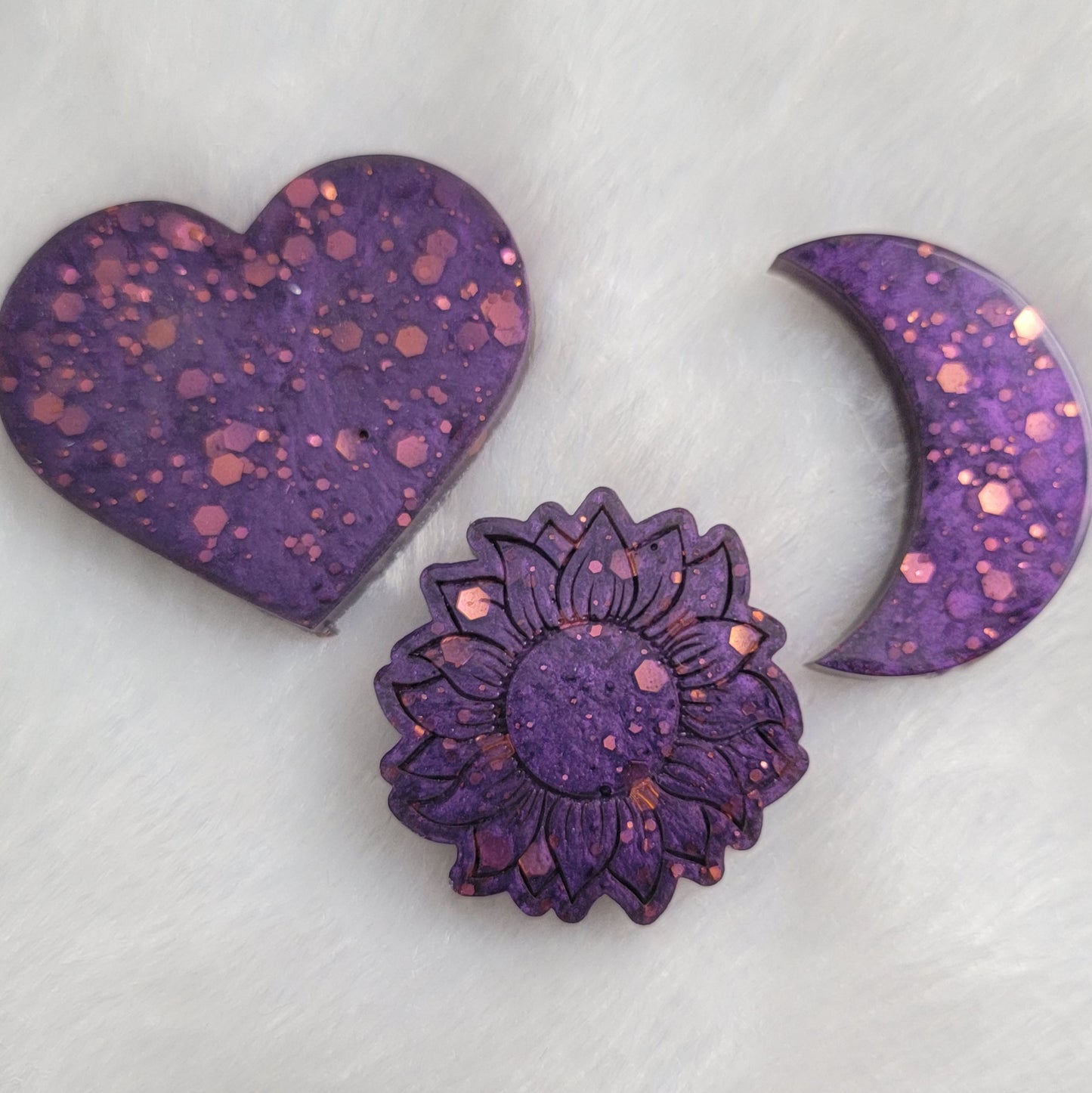 Three epoxy resin pieces - heart, moon, and flower shaped - made with pink glitter and purple pigment.