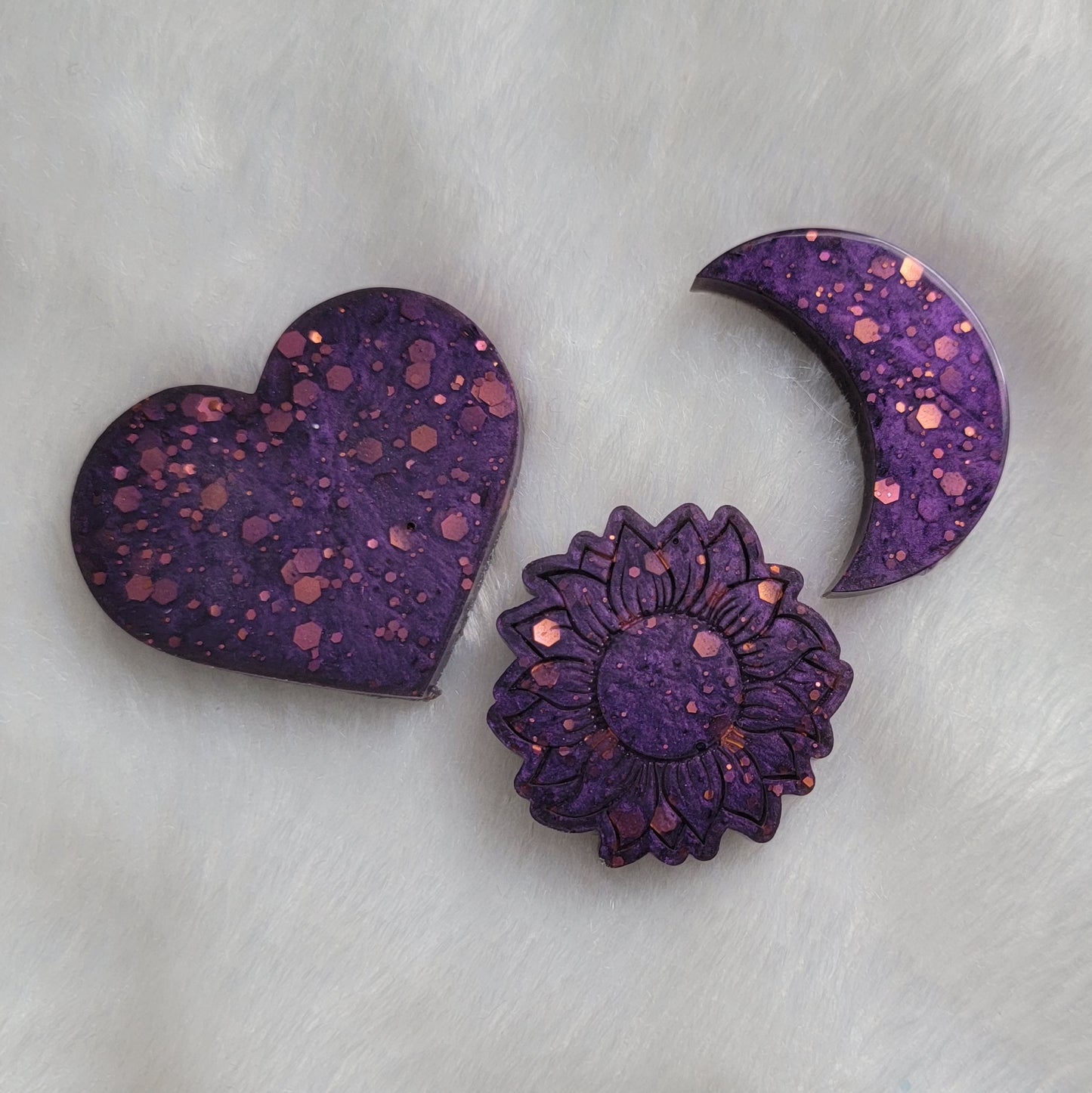 Three epoxy resin pieces - heart, moon, and flower shaped - made with pink glitter and purple pigment.