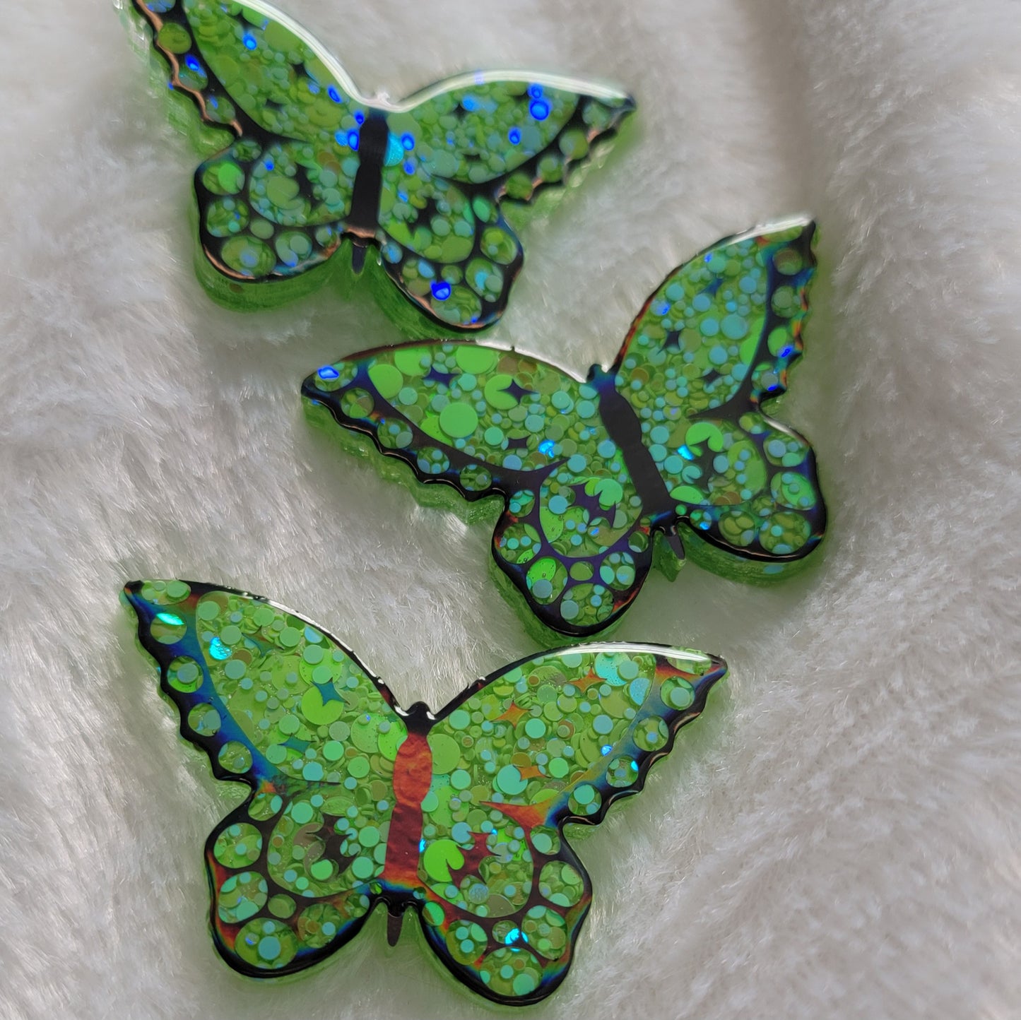 Three butterfly shaped epoxy resin pieces, made with bright green glitter and holographic black vinyl details.