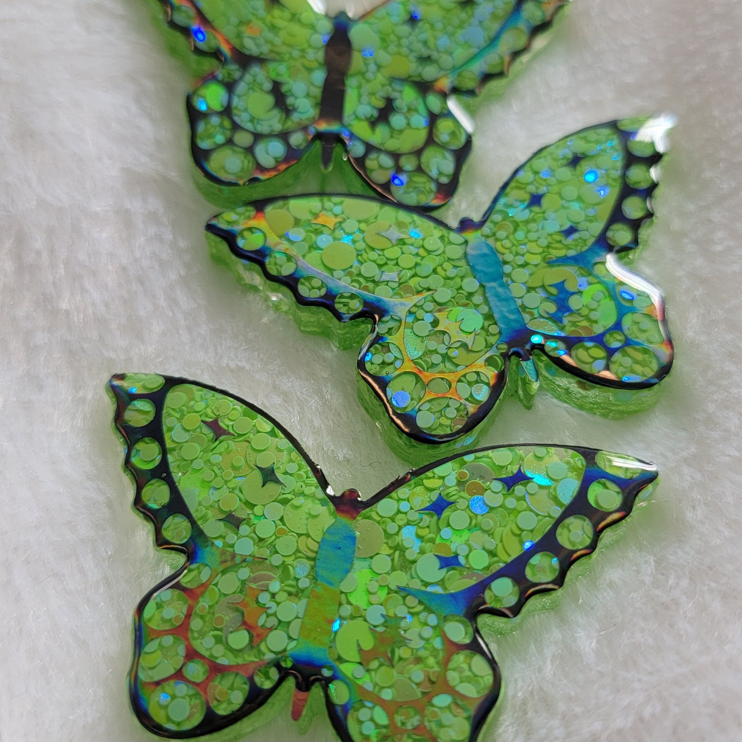Three butterfly shaped epoxy resin pieces, made with bright green glitter and holographic black vinyl details.