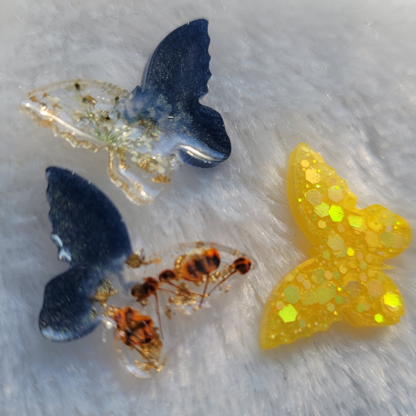 Three extra small butterfly shaped epoxy resin pieces, one with bright yellow glitter and two with dark silver blue pigment and pressed flowers - one orange and one white.