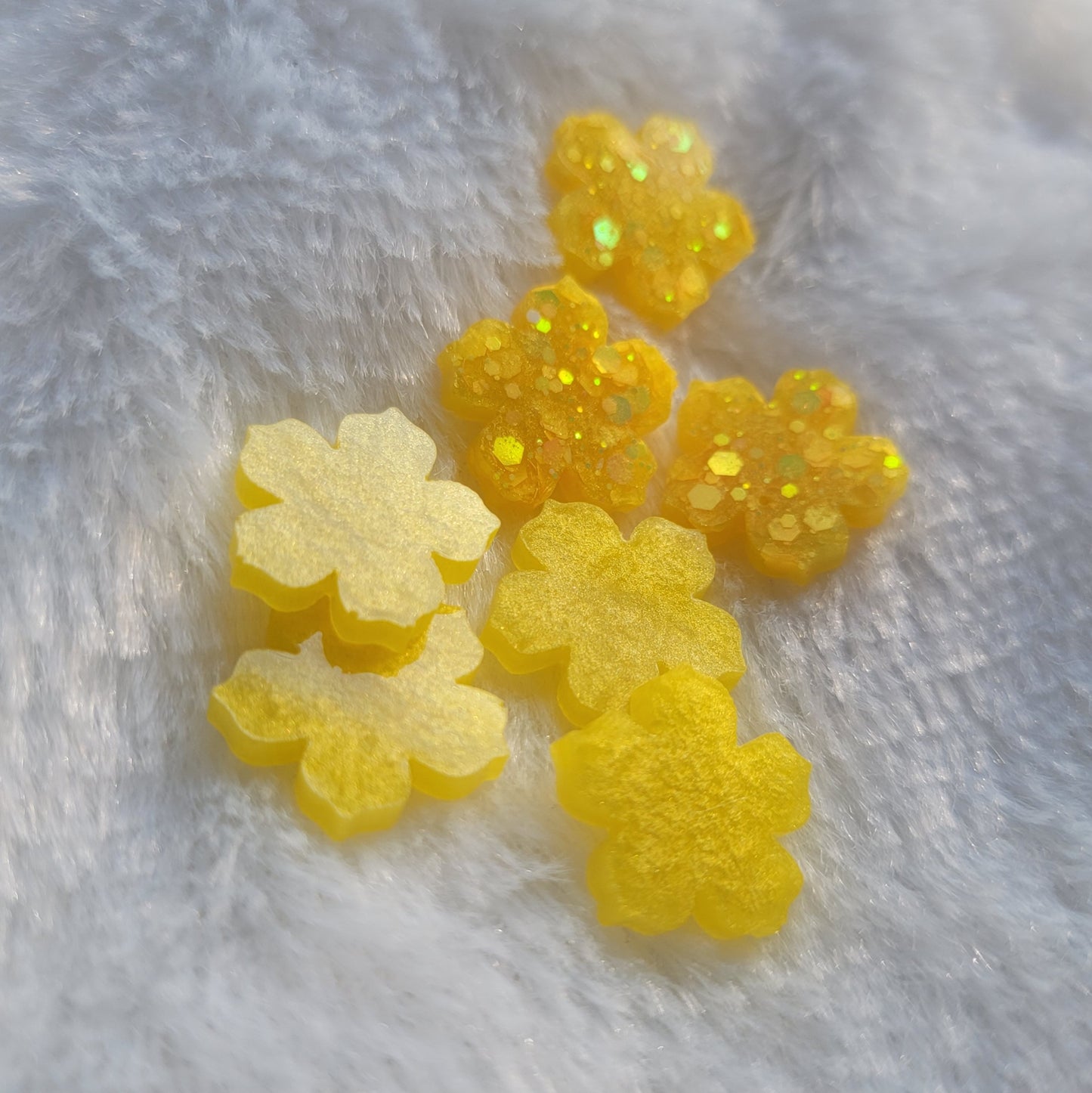 Seven extra small flower blossom shaped epoxy resin pieces, four with shimmering yellow pigment and three with bright yellow glitter.