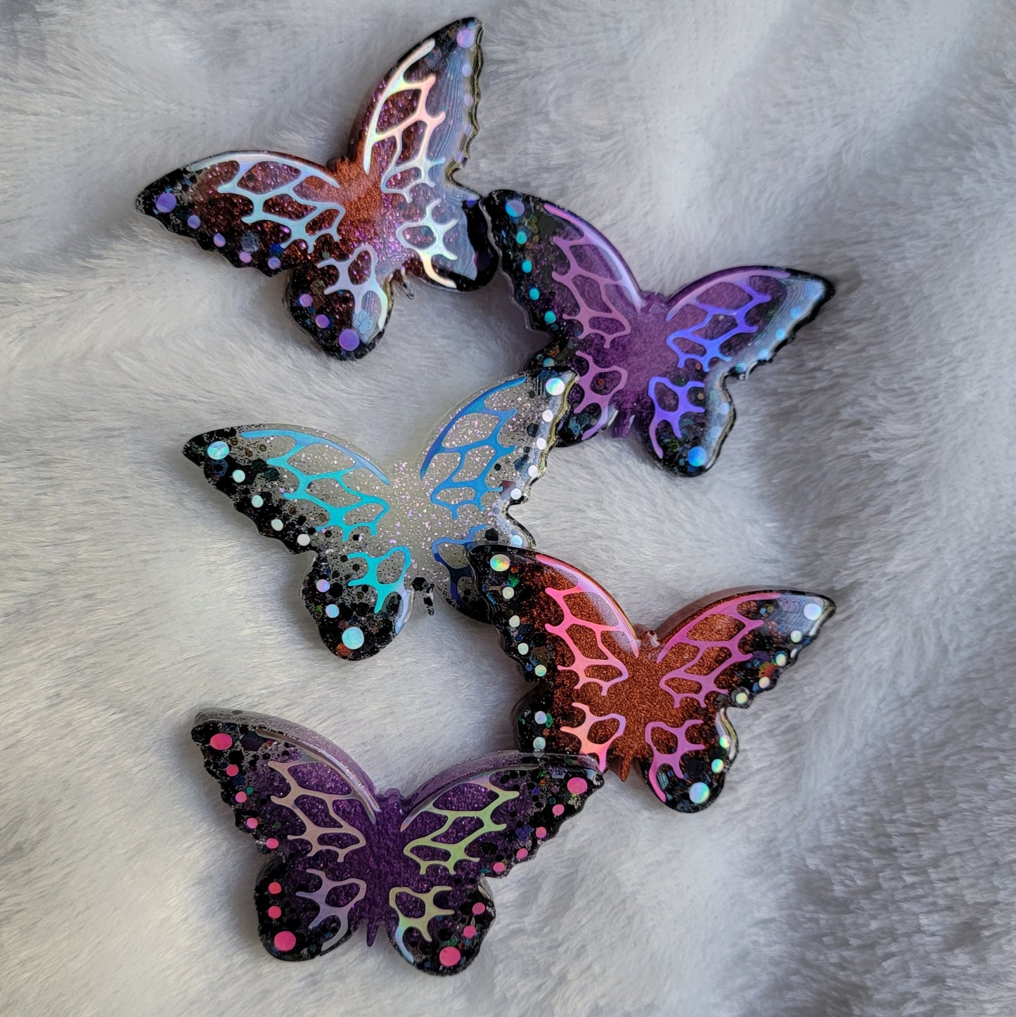 Five butterfly shaped epoxy resin pieces with purple, copper, and white bases and holographic black glitter edges. Each decorated with intricate holographic vinyl wing veins and dots in unique colors.