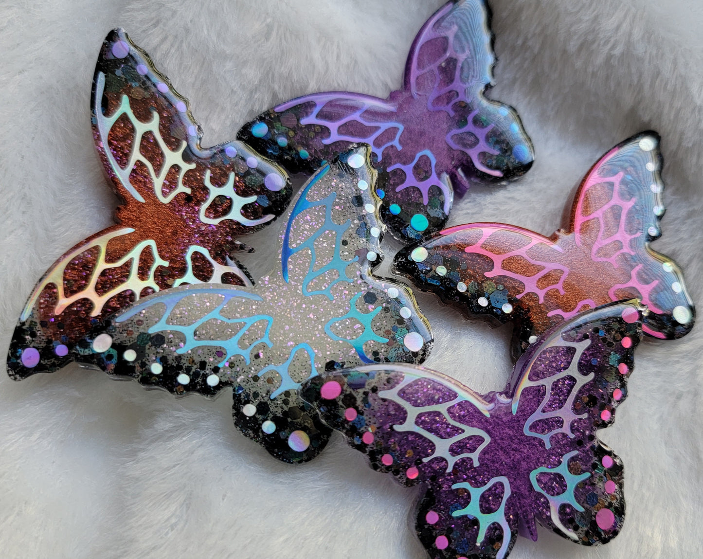 Five butterfly shaped epoxy resin pieces with purple, copper, and white bases and holographic black glitter edges. Each decorated with intricate holographic vinyl wing veins and dots in unique colors.