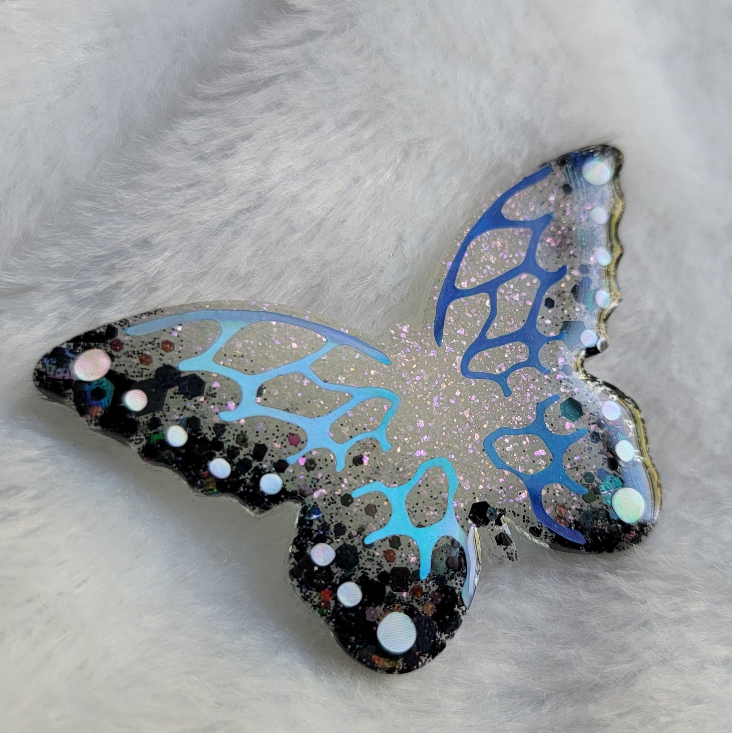 Butterfly shaped epoxy resin piece, shimmering white and black glitter base decorated with holographic blue and silver vinyl.