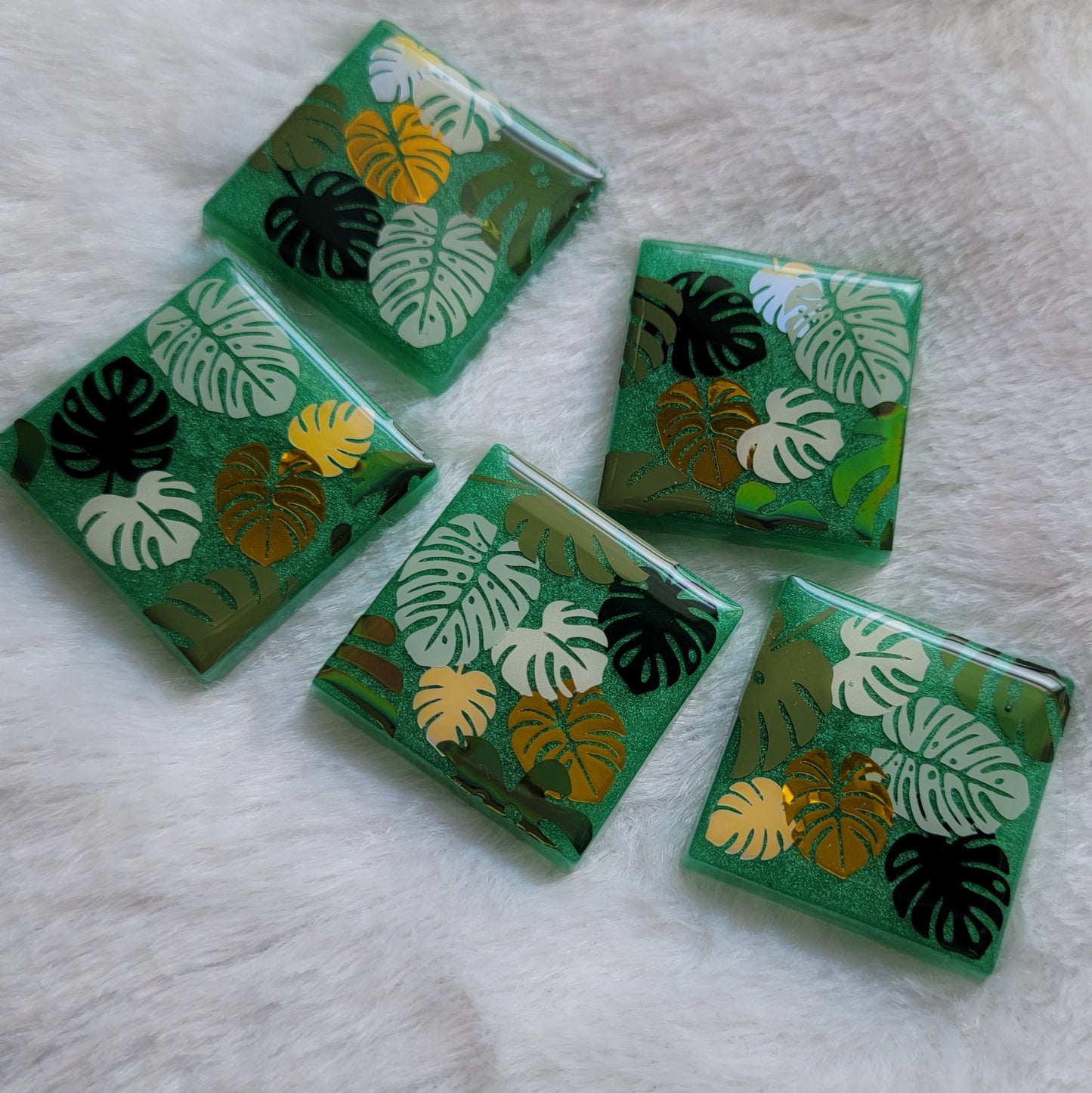Five square epoxy resin pieces with emerald green bases, each decorated with a unique monstera leaf vinyl design in shades of green and gold.
