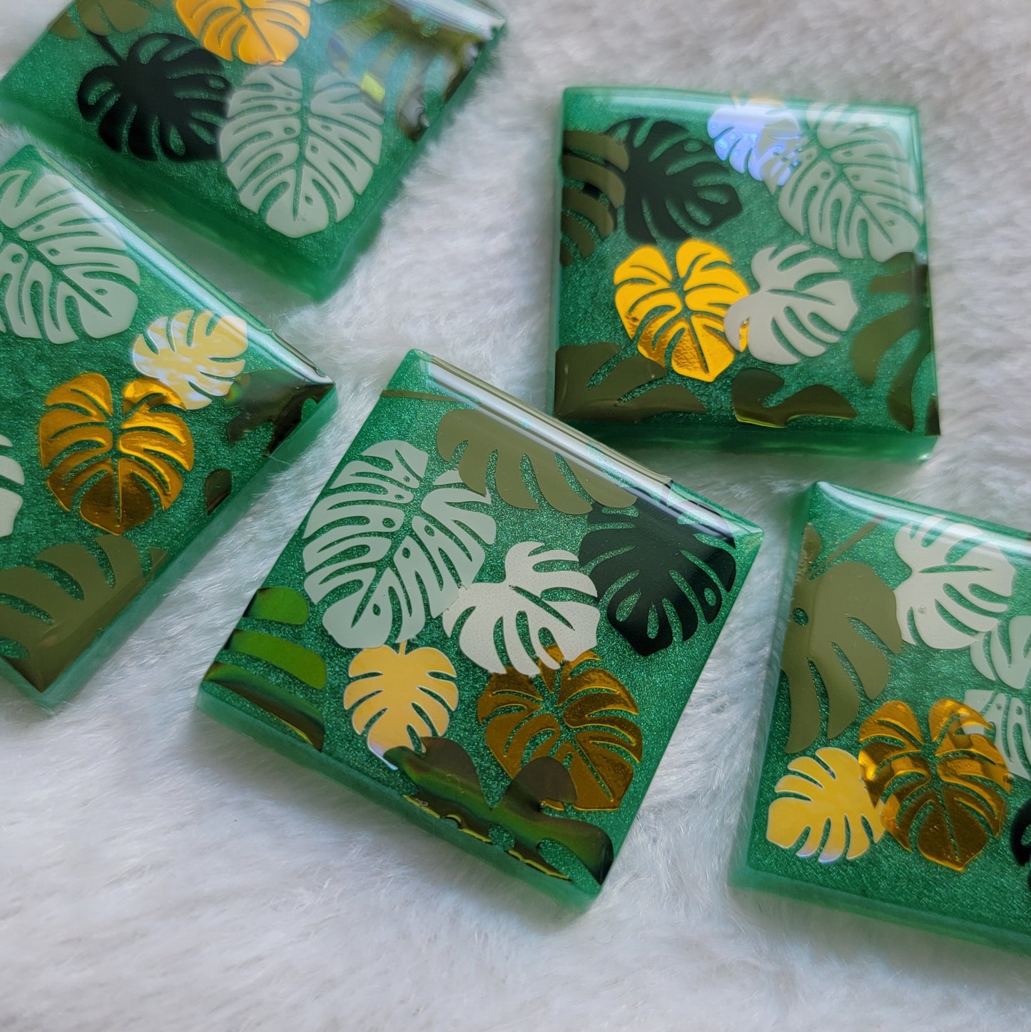 Five square epoxy resin pieces with emerald green bases, each decorated with a unique monstera leaf vinyl design in shades of green and gold.