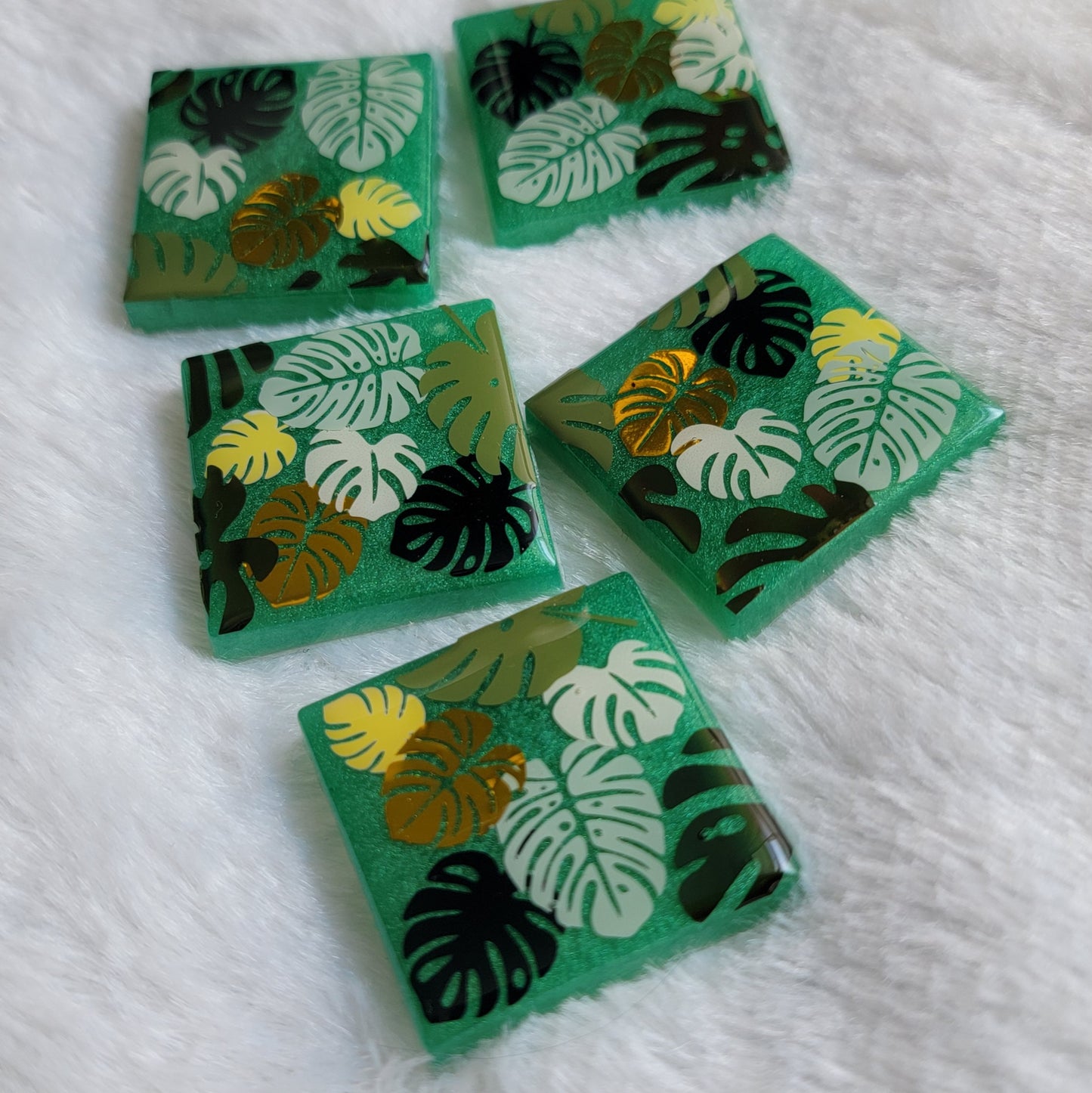 Five square epoxy resin pieces with emerald green bases, each decorated with a unique monstera leaf vinyl design in shades of green and gold.