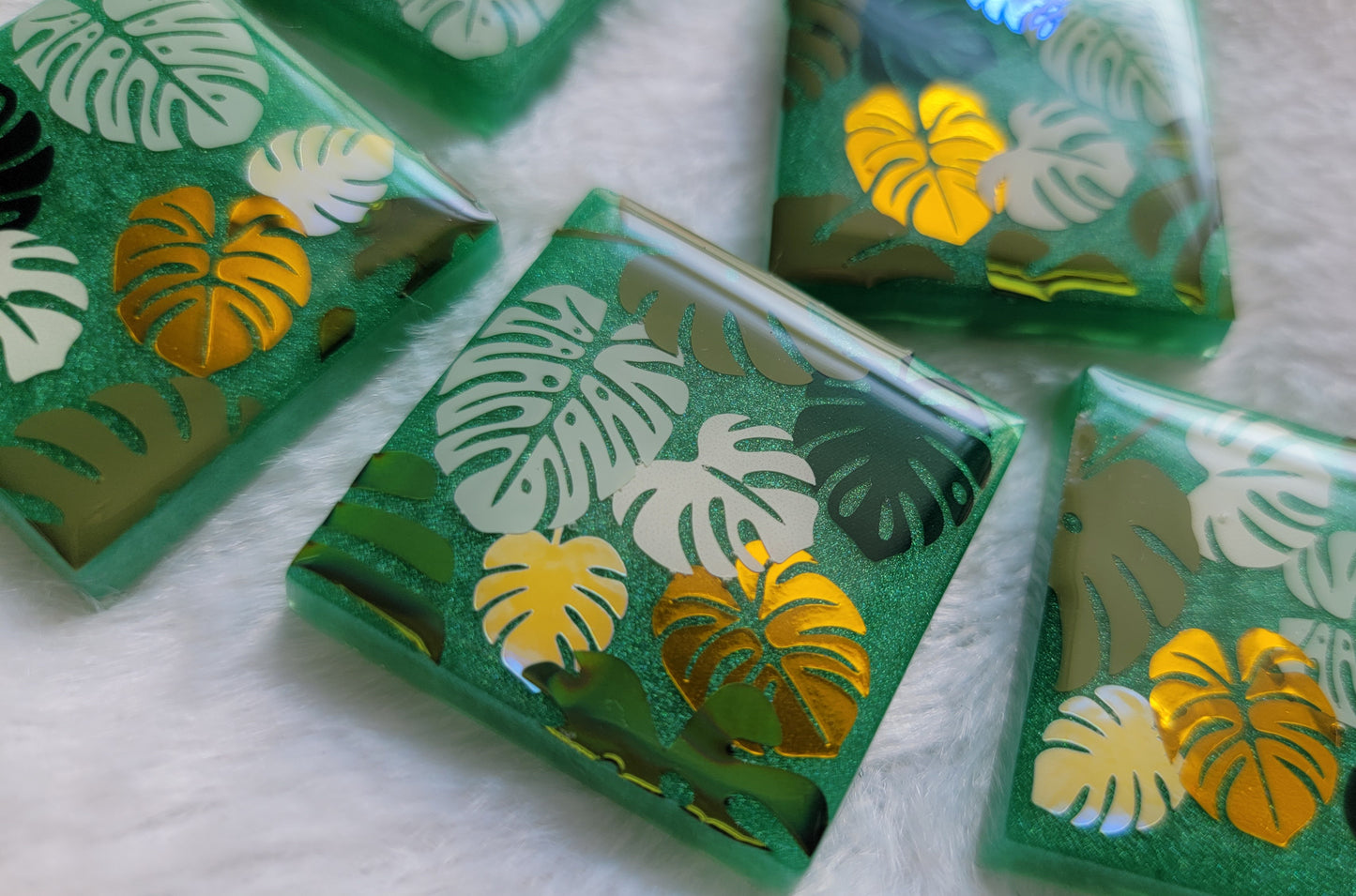 Close up photo of five square epoxy resin pieces with emerald green bases, each decorated with a unique monstera leaf vinyl design in shades of green and gold.