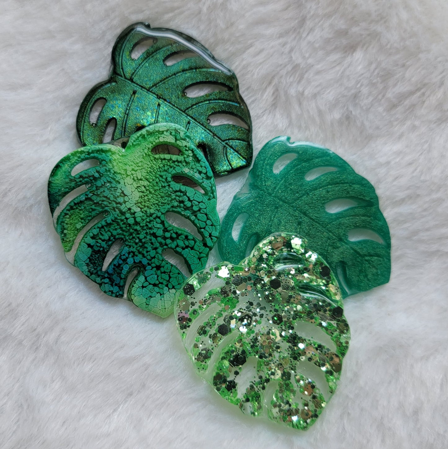 Four epoxy resin pieces shaped like monstera leaves in four different color options - chameleon green, green ink, emerald green, light green glitter.