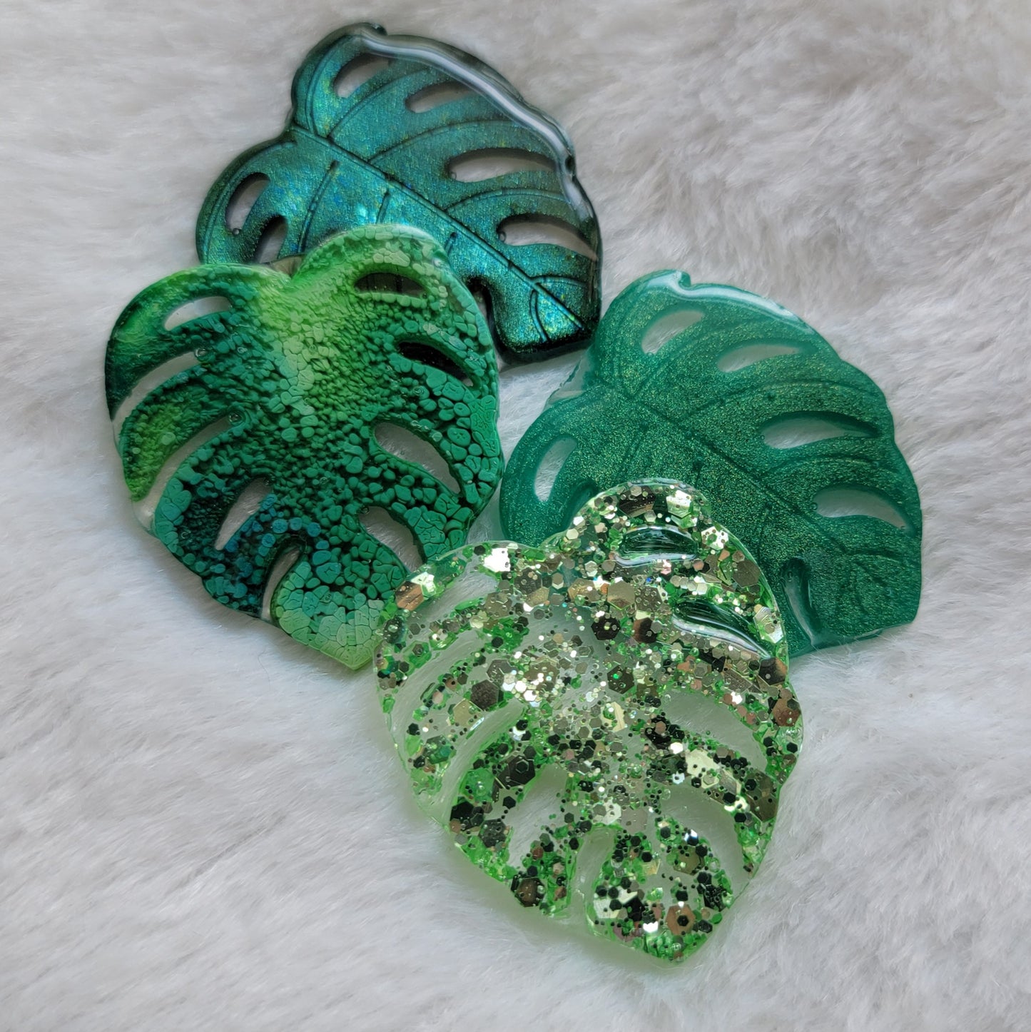 Four epoxy resin pieces shaped like monstera leaves in four different color options - chameleon green, green ink, emerald green, light green glitter.