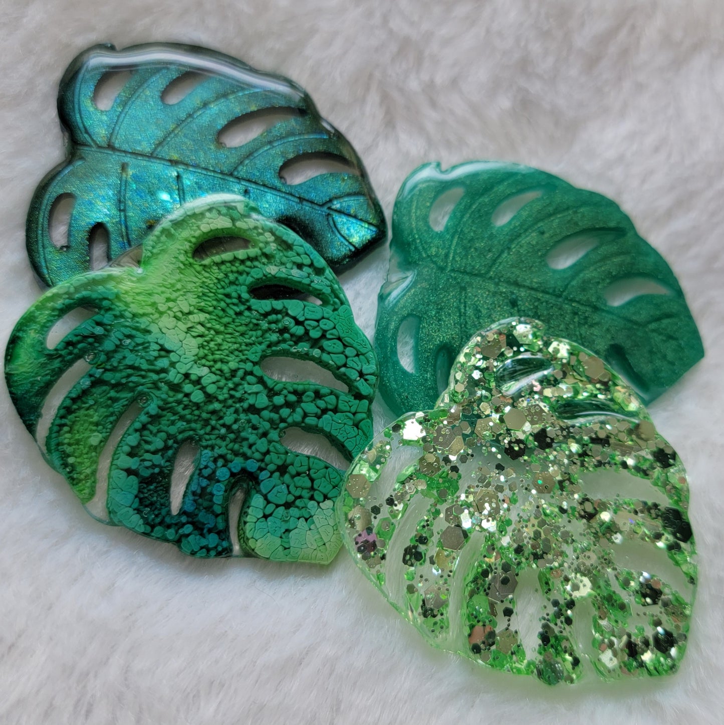 Four epoxy resin pieces shaped like monstera leaves in four different color options - chameleon green, green ink, emerald green, light green glitter.