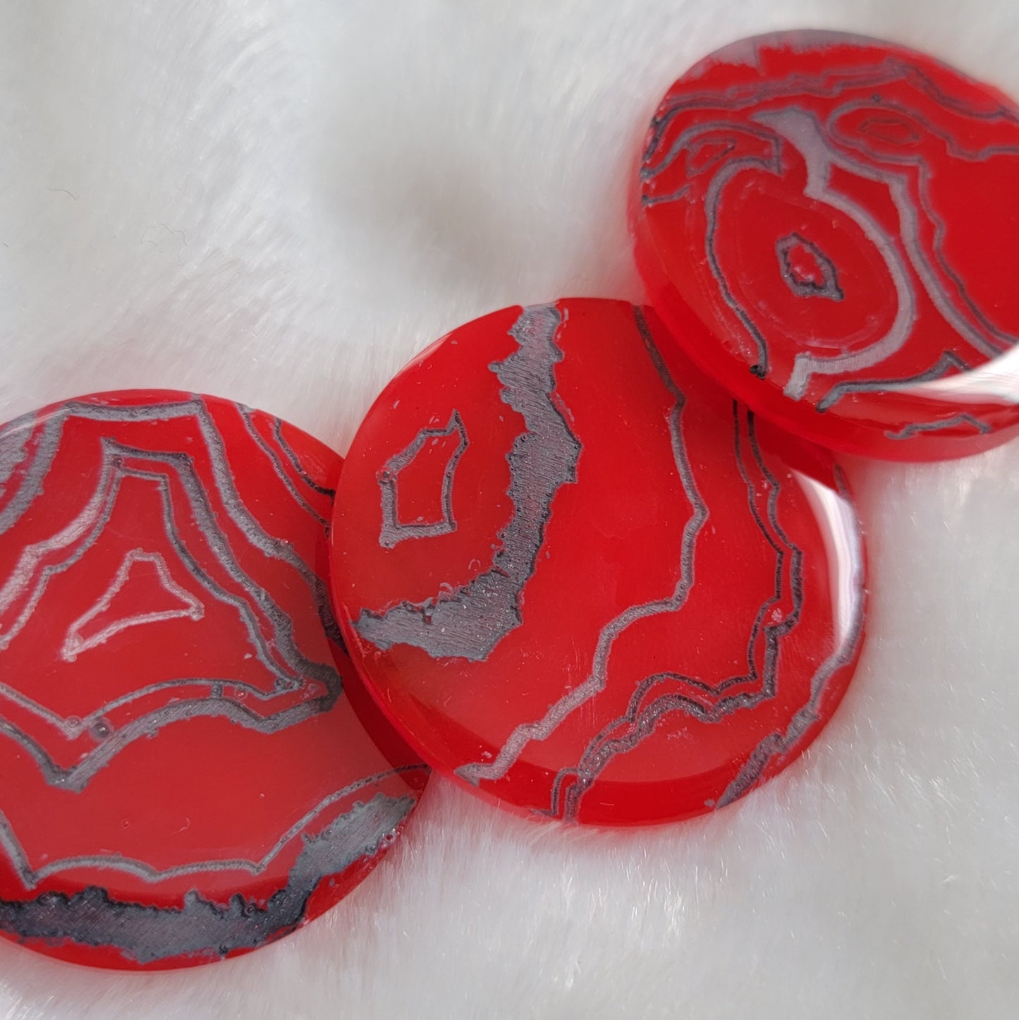 Three bright red slightly translucent round epoxy resin pieces with engraved details that resemble geode layers in shades of silver and grey.