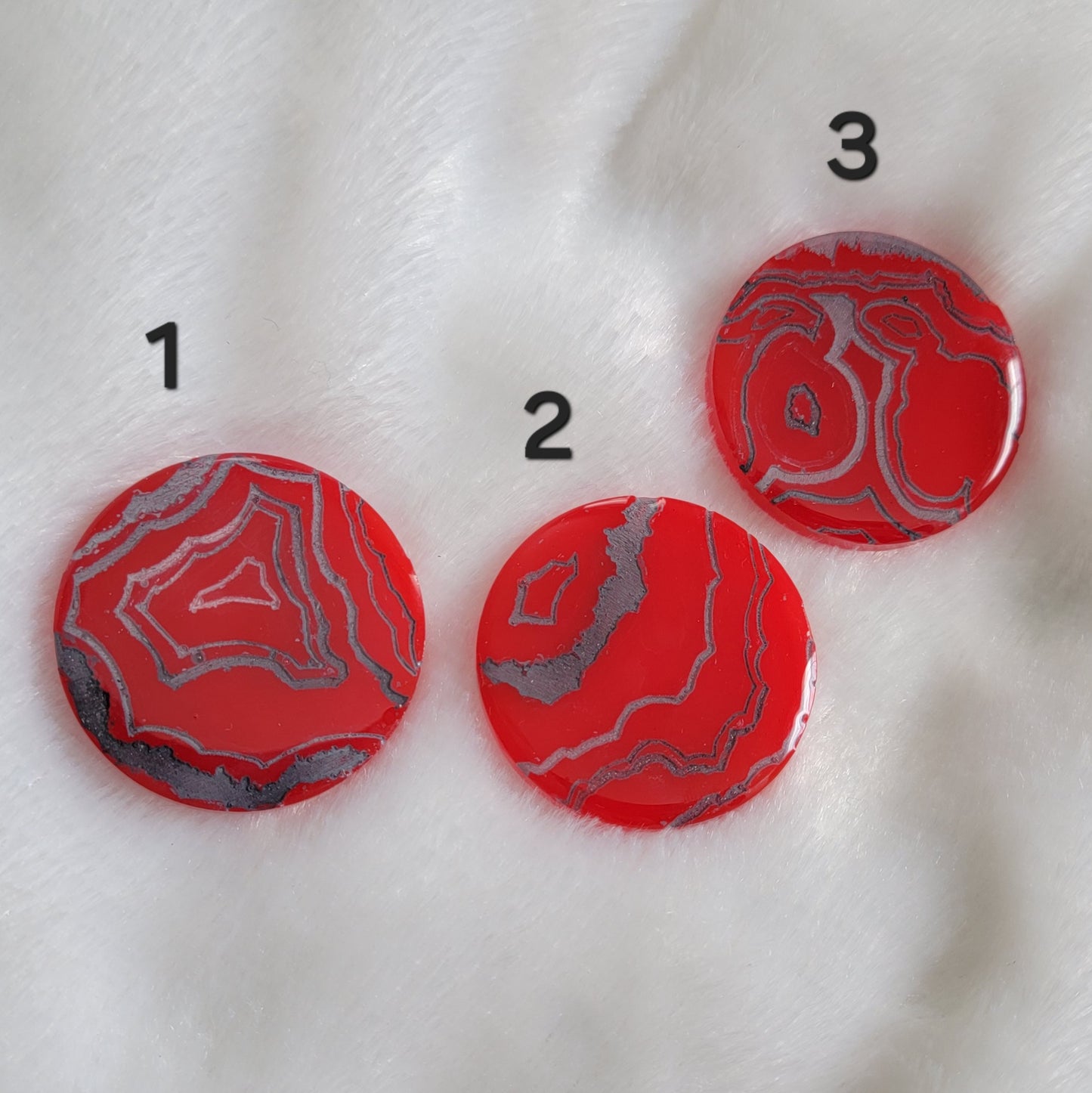 Three bright red slightly translucent round epoxy resin pieces with engraved details that resemble geode layers in shades of silver and grey.