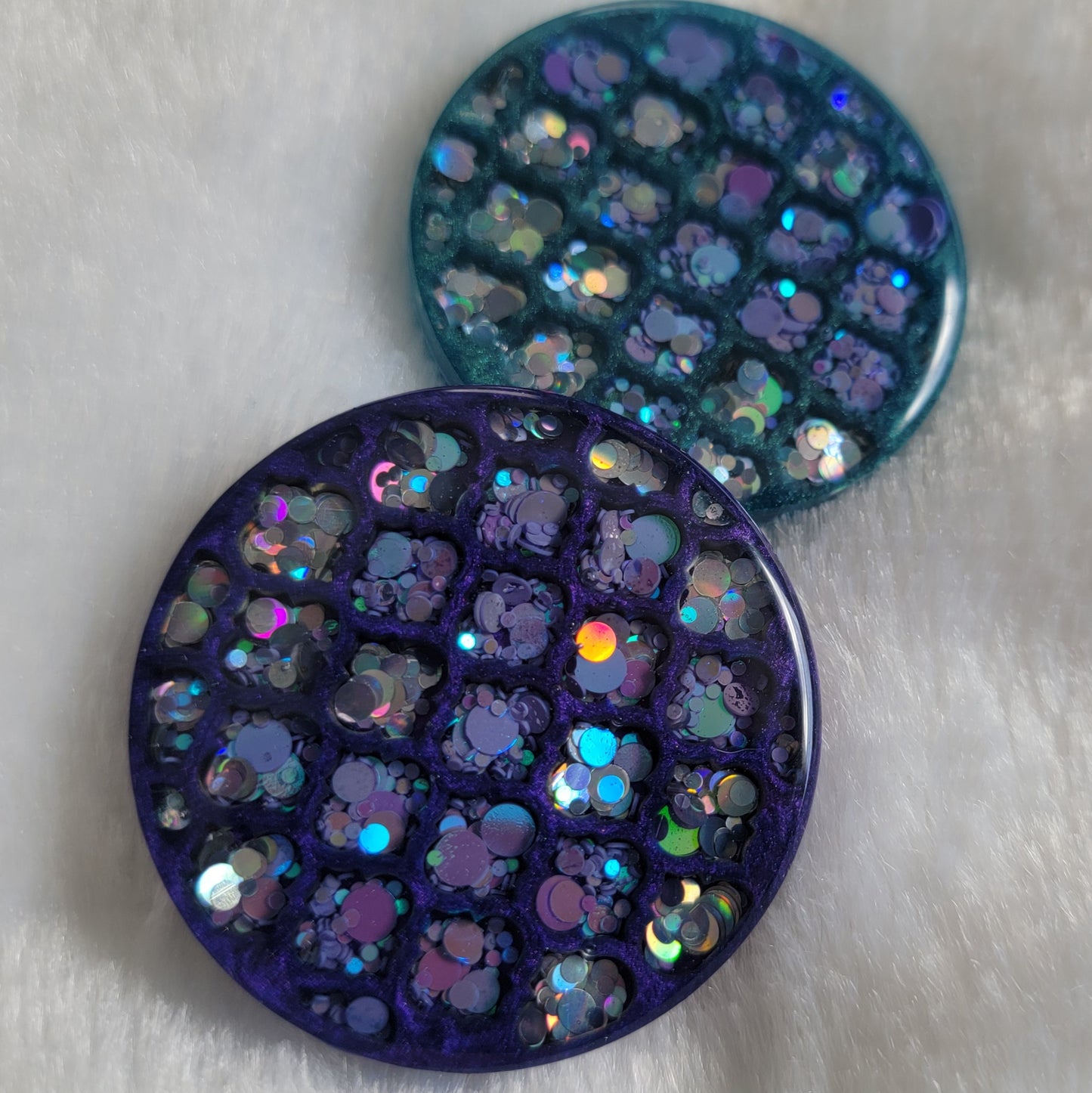 Two round epoxy resin pieces with geometric lattice framework in dark purple and teal, accented with silver and lavender holographic glitter.