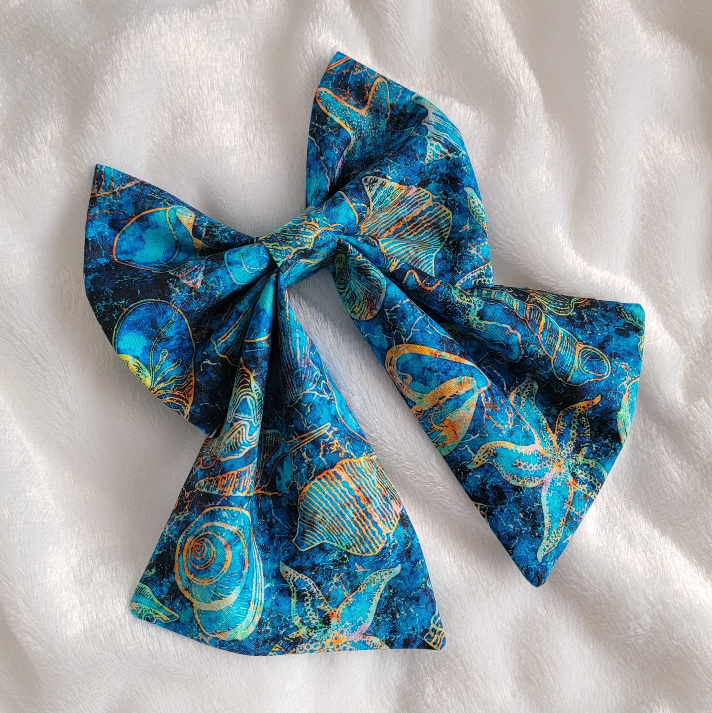 A handmade pet bow with a bold cotton print in ultramarine blue with amber outlines of seashells and starfish, sailor bow style with long showy tails.