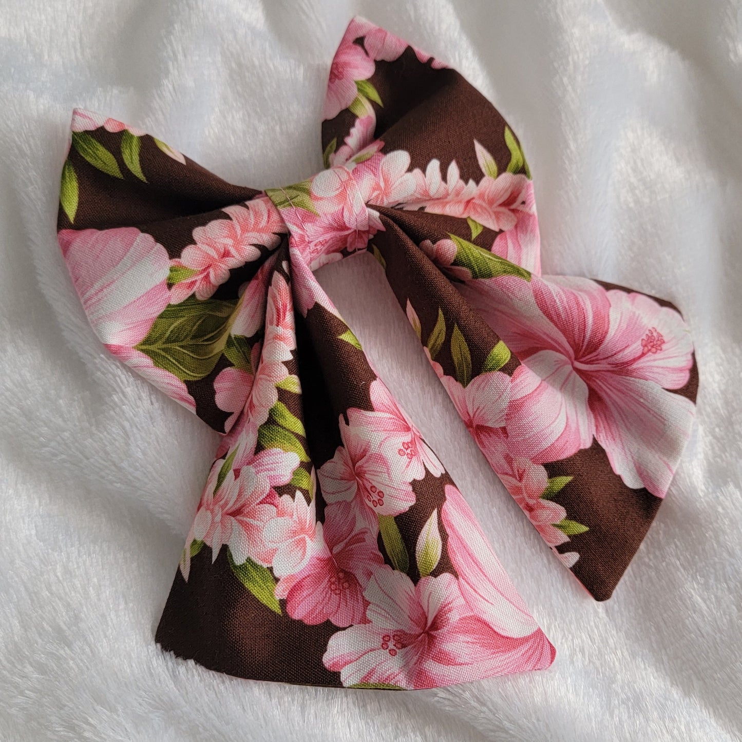 A fabric pet bow with a floral pattern of pink hibiscus leis on rich brown background, sailor bow style with long showy tails.