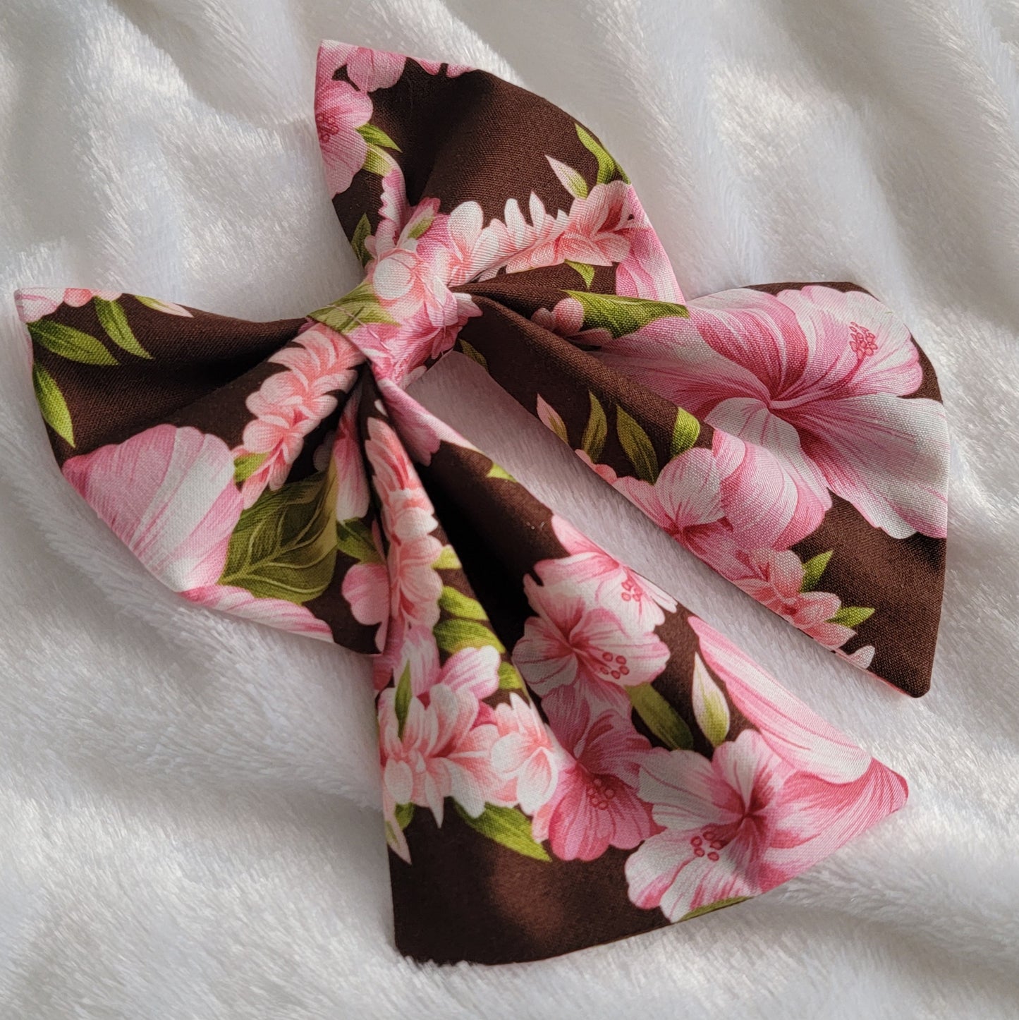 A fabric pet bow with a floral pattern of pink hibiscus leis on rich brown background, sailor bow style with long showy tails.