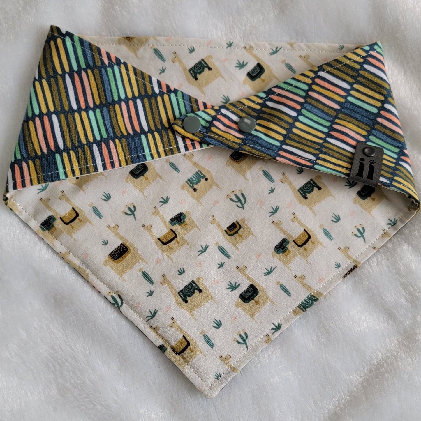 A reversible pet bandana with one side featuring llamas and cactus on a beige background and the other side with stripes in various earthy colors.