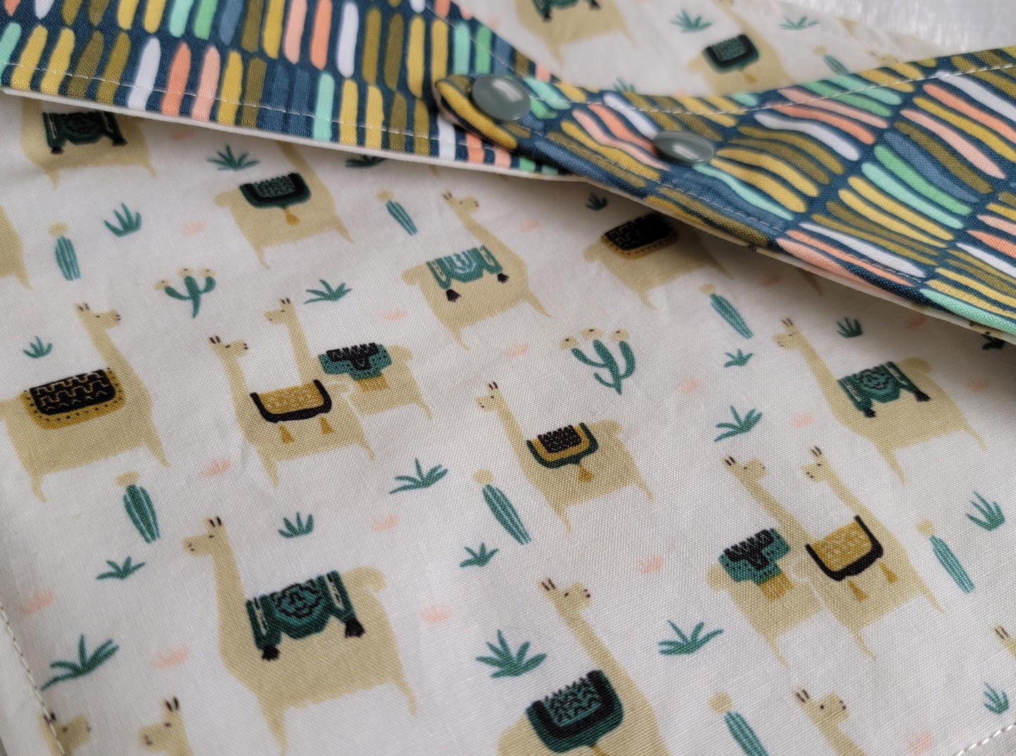 A reversible pet bandana with one side featuring llamas and cactus on a beige background and the other side with stripes in various earthy colors.