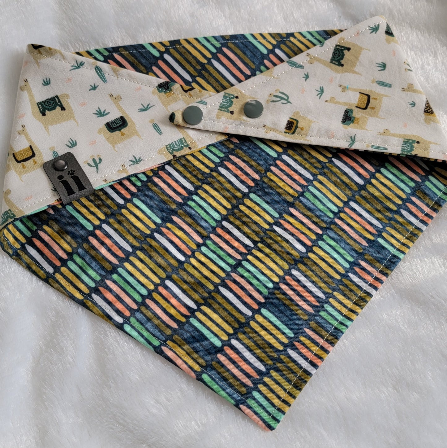 A reversible pet bandana with one side featuring llamas and cactus on a beige background and the other side with stripes in various earthy colors.