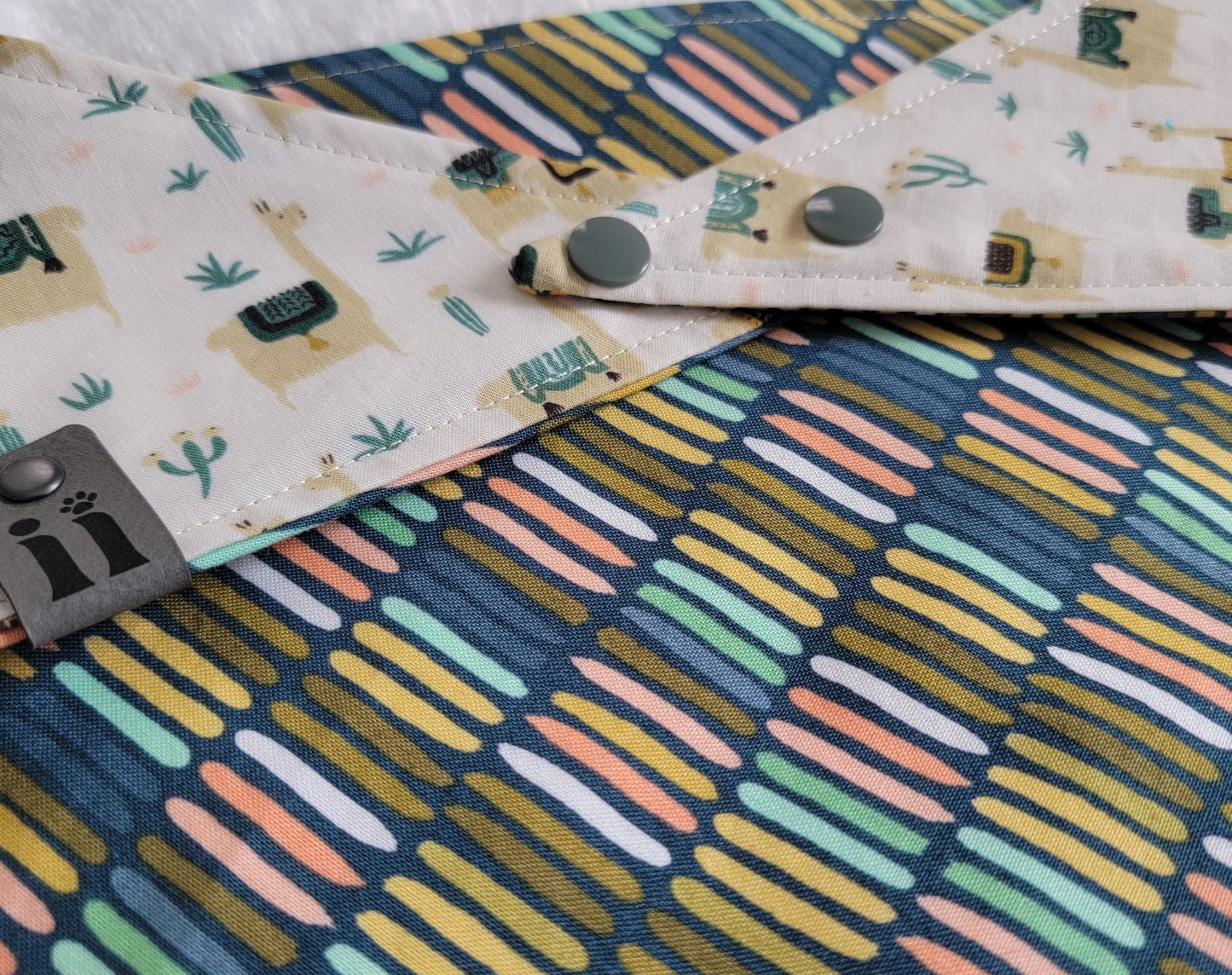 A reversible pet bandana with one side featuring llamas and cactus on a beige background and the other side with stripes in various earthy colors.