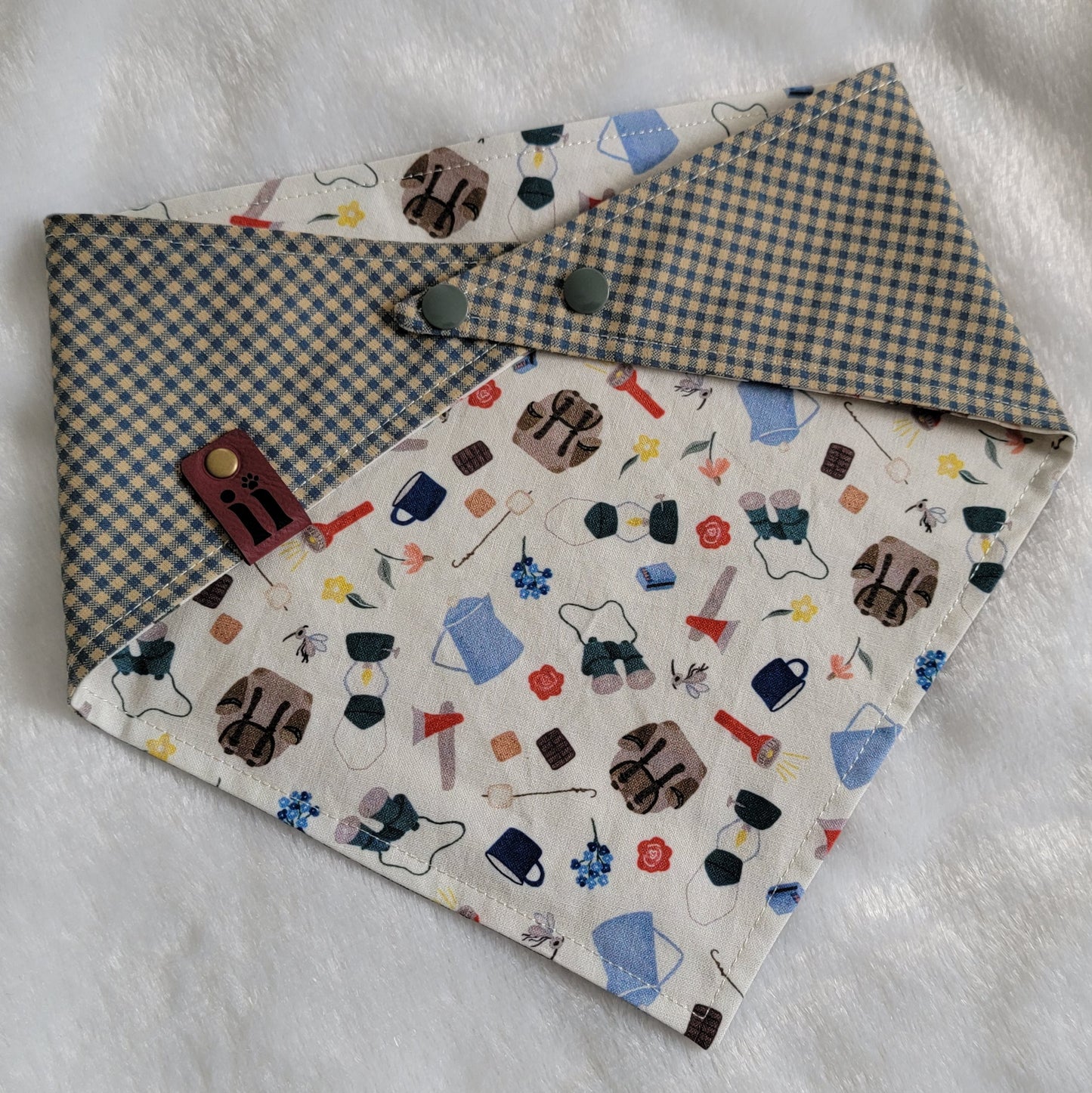 A reversible pet bandana with one side featuring an assortment of camping-themed patterns including a tent, campfire, and backpack, and the other side in a blue gingham plaid.