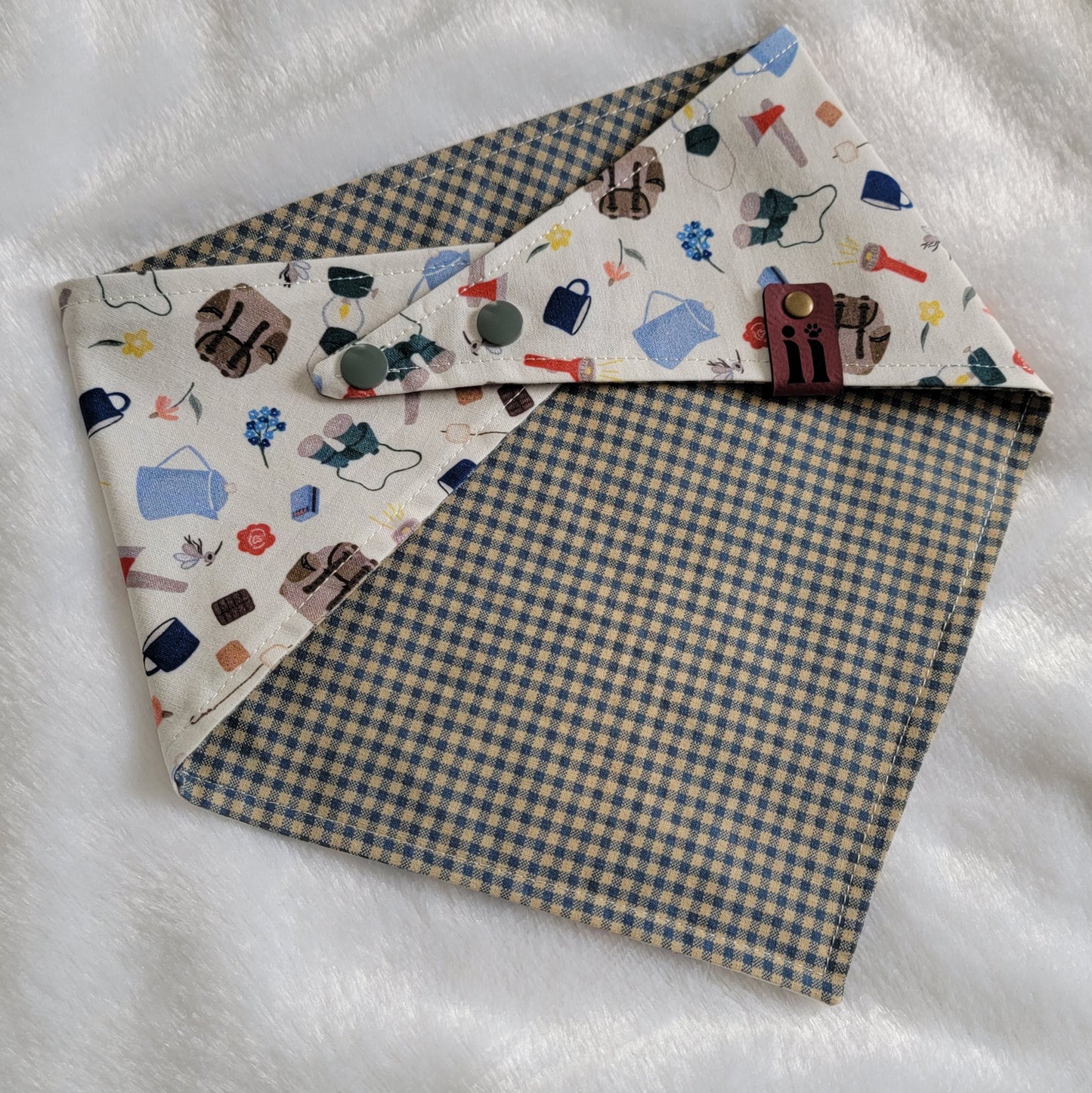 A reversible pet bandana with one side featuring an assortment of camping-themed patterns including a tent, campfire, and backpack, and the other side in a blue gingham plaid.