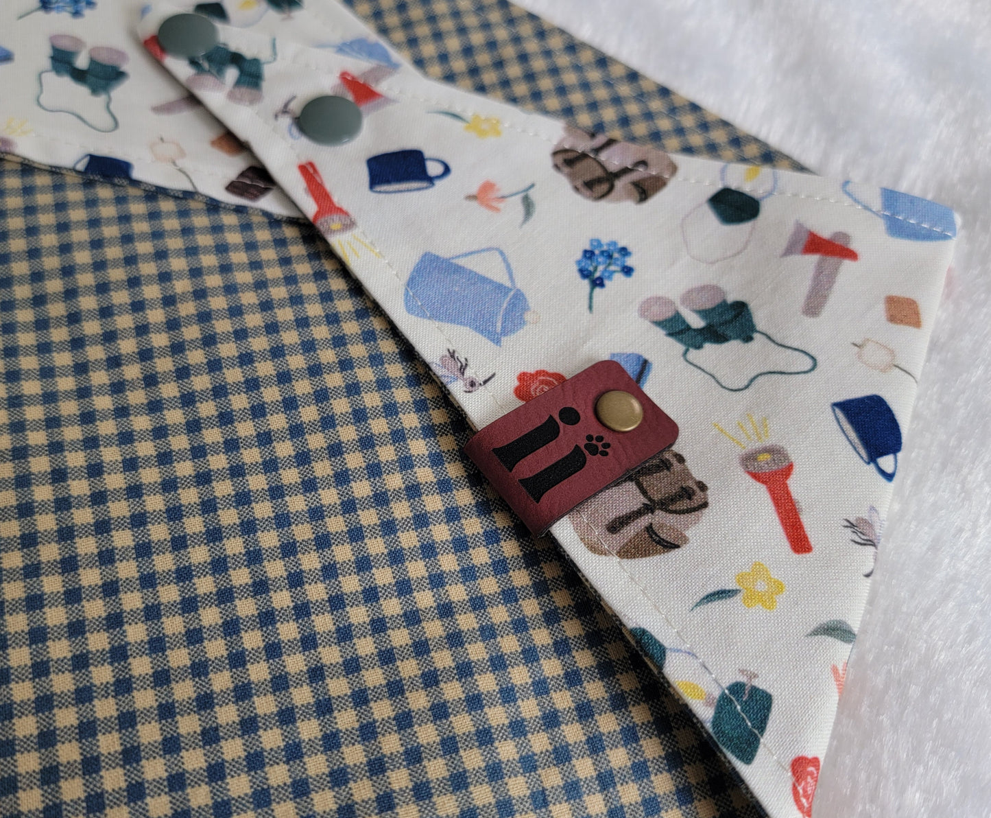 A reversible pet bandana with one side featuring an assortment of camping-themed patterns including a tent, campfire, and backpack, and the other side in a blue gingham plaid.