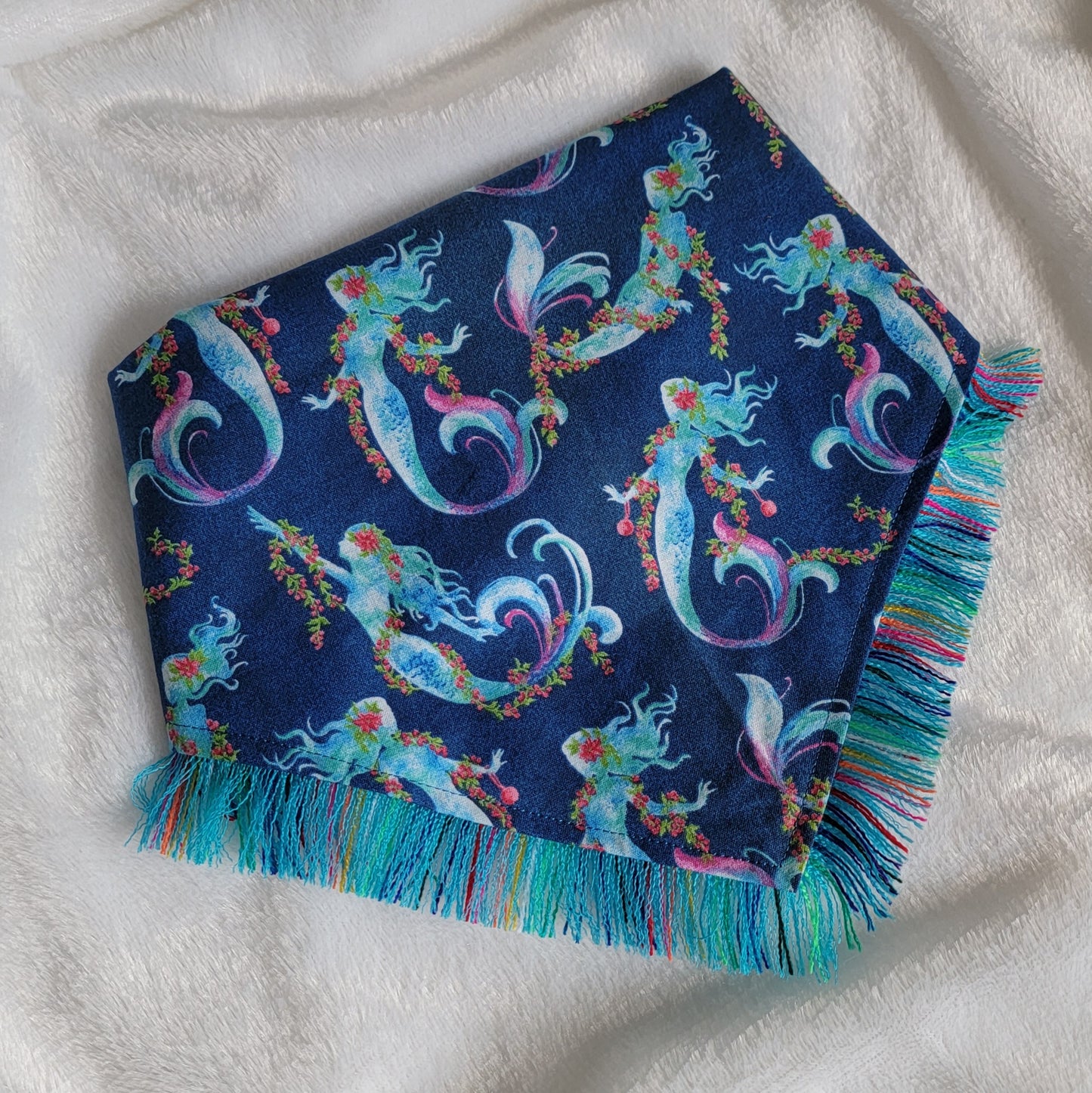 A blue bandana with a mermaid pattern and vibrant blue and rainbow fringe edges.