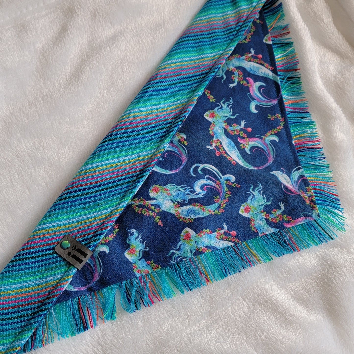 A blue bandana with a mermaid pattern and vibrant blue and rainbow fringe edges.