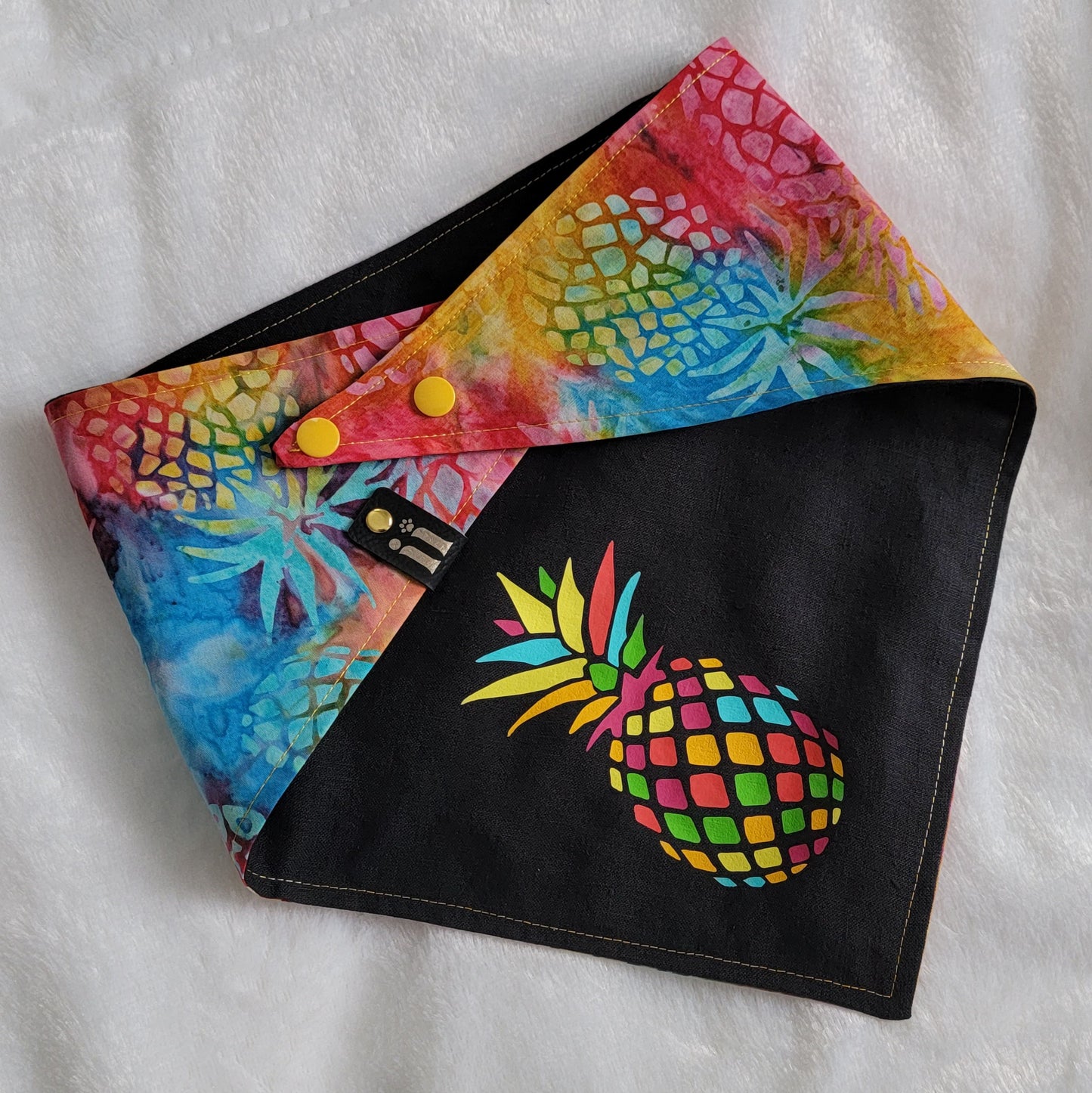 A reversible pet bandana with a vibrant rainbow pineapple batik pattern on one side and a solid black hemp cotton blend with a custom vinyl pineapple design on the other, displayed on a white background.