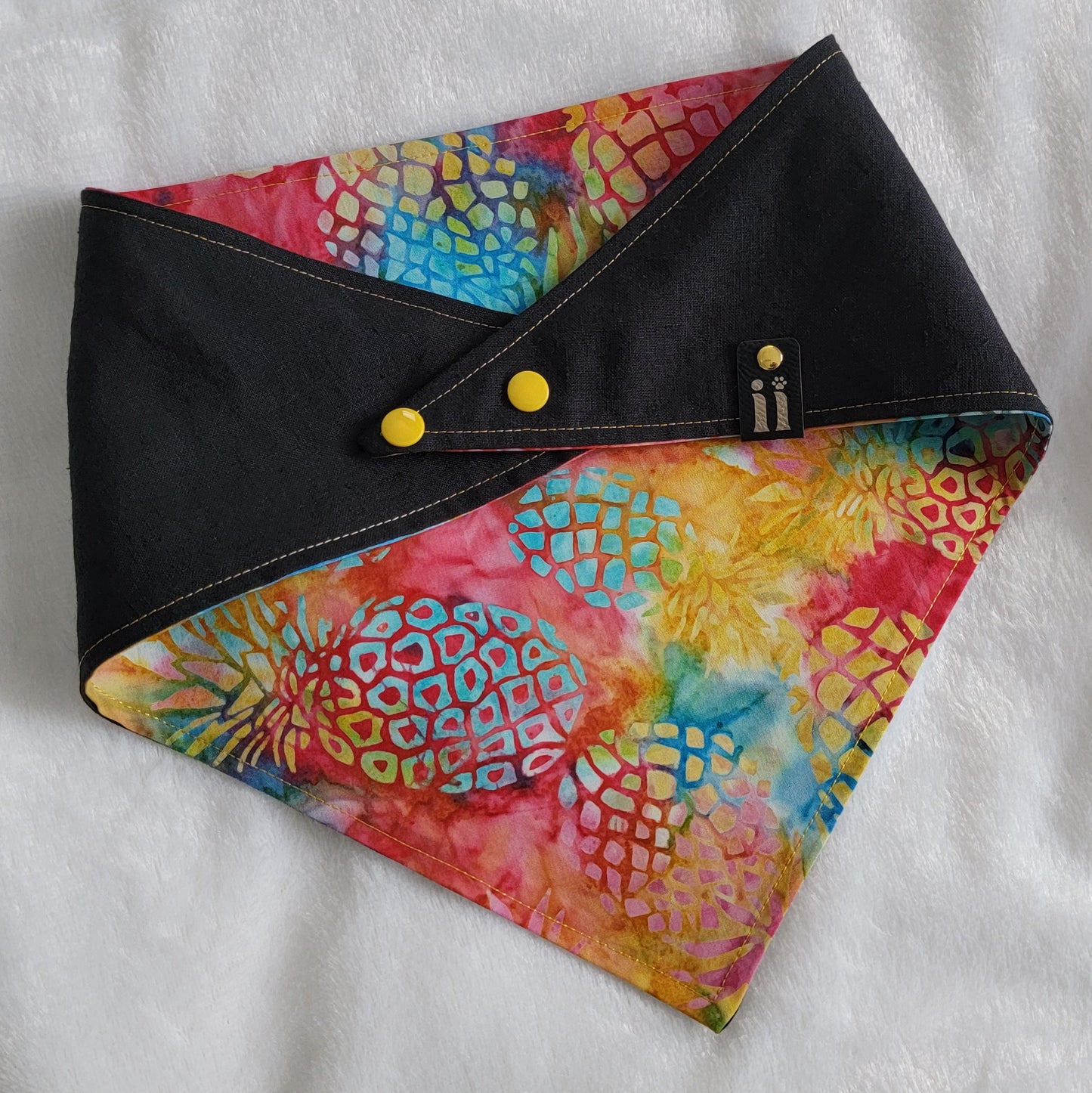 A reversible pet bandana with a vibrant rainbow pineapple batik pattern on one side and a solid black hemp cotton blend with a custom vinyl pineapple design on the other, displayed on a white background.