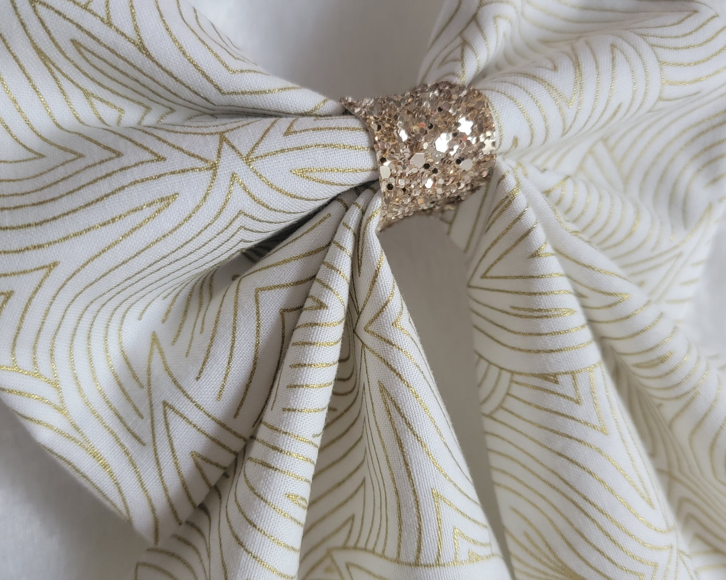 Close up of handmade pet bow with a soft cotton print of abstract gold geometric lines on off-white background, gold blue glitter center sailor bow style with long showy tails.
