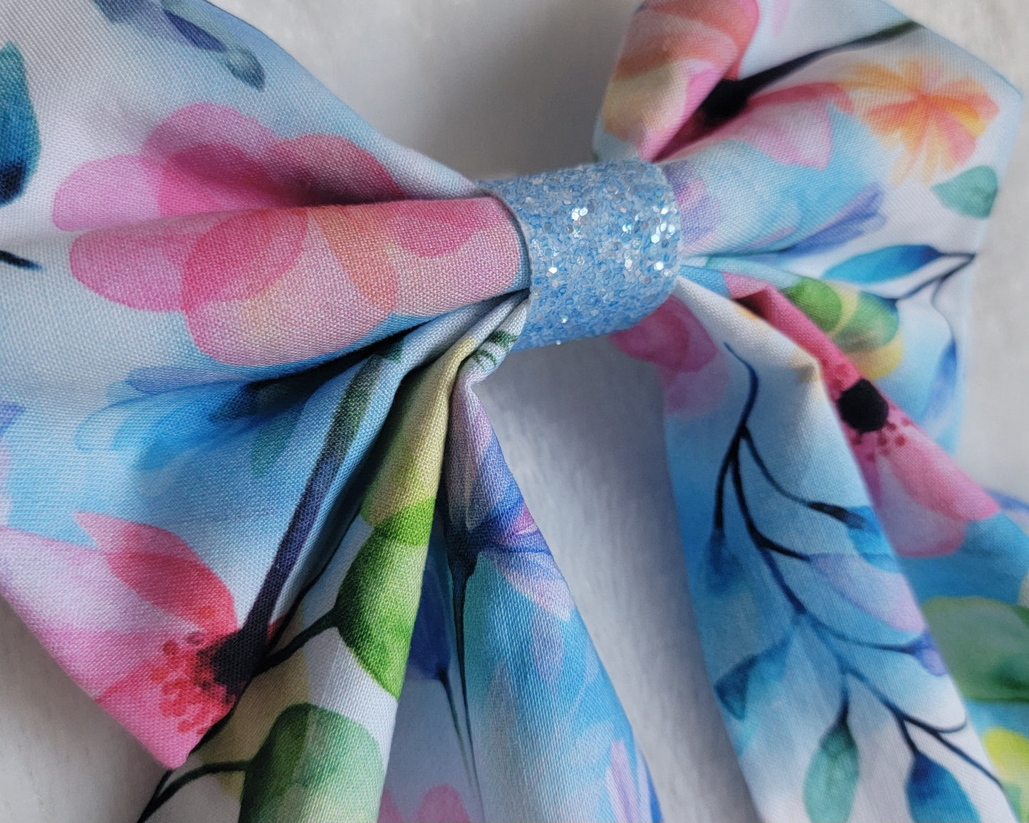 Close up of handmade pet bow with a soft cotton print of watercolor florals in light blues pinks and greens, light blue glitter center sailor bow style with long showy tails.
