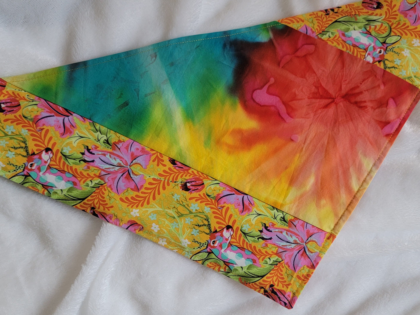 A multicolored pet bandana with stripes of cotton fabric featuring colorful rainbow tie-dye and vibrant floral stag print. Laying flat showing side that has narrow edge stripe of stag floral fabric.