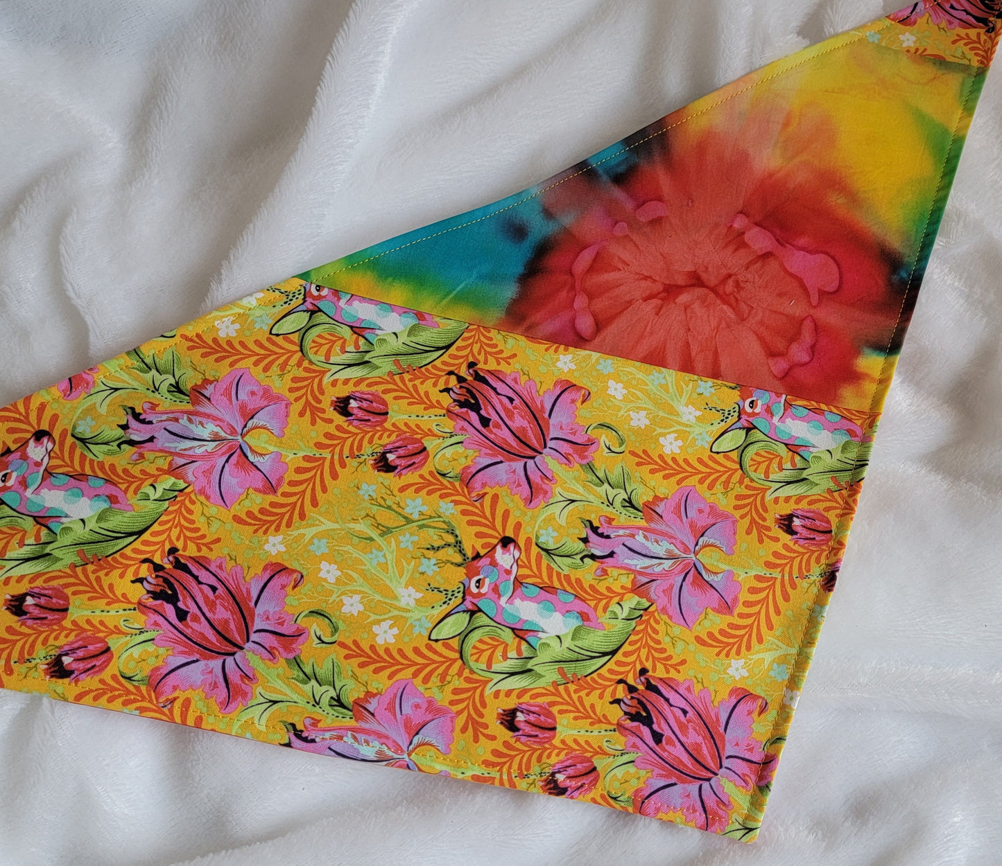 A multicolored pet bandana with stripes of cotton fabric featuring colorful rainbow tie-dye and vibrant floral stag print. Laying flat showing side that has wider stripe of stag floral fabric.