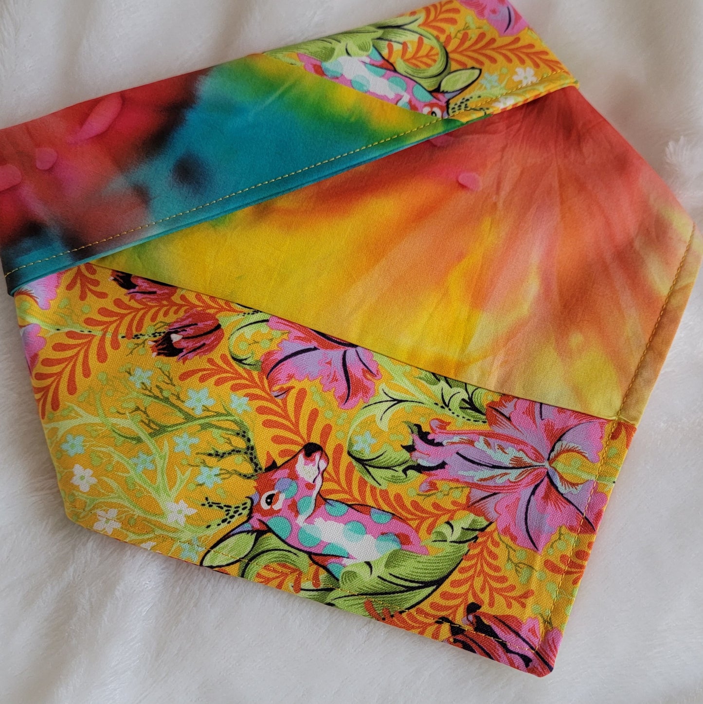 A multicolored pet bandana with stripes of cotton fabric featuring colorful rainbow tie-dye and vibrant floral stag print. Showing side that has narrower edge stripe of stag floral fabric with folded corners and top to show edge or reverse side.