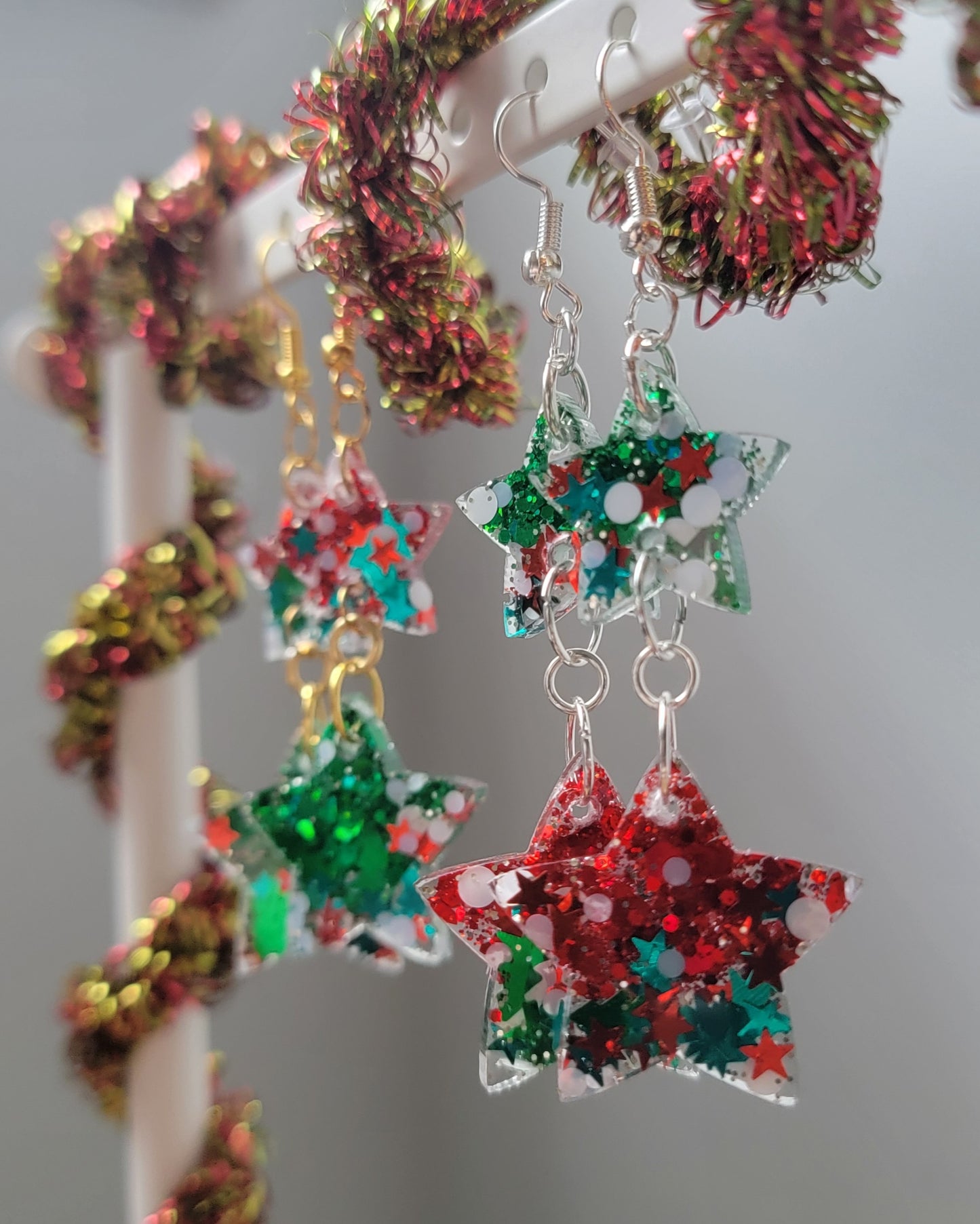 Two pairs of drop earrings, green and red and Christmas glitters in star shaped resin pieces with gold colored and silver colored metal findings. Earrings are displayed on a white earring hanger with red and gold garland wrapped around it.