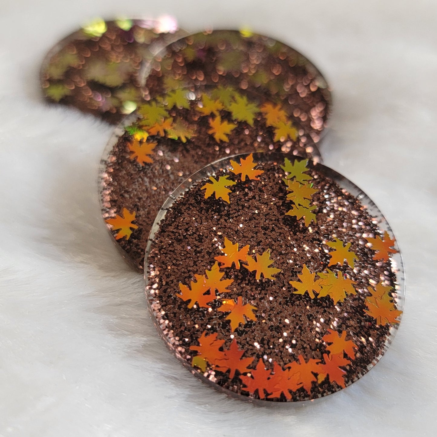 Four round epoxy resin pieces with dark brown glitter and holographic orange fall leaf shaped glitter.