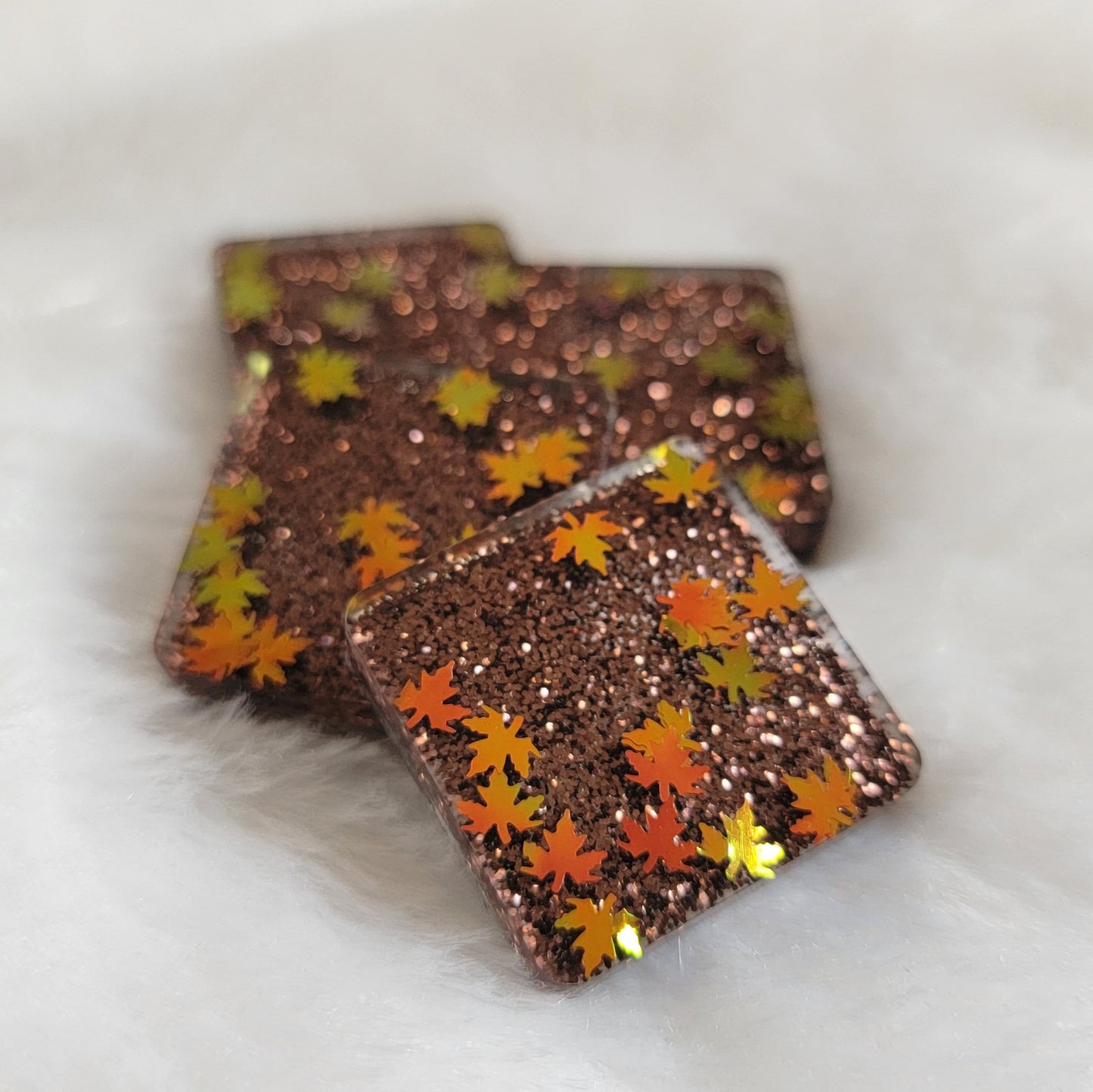 Four square poxy resin pieces with dark brown glitter and holographic orange fall leaf shaped glitter.