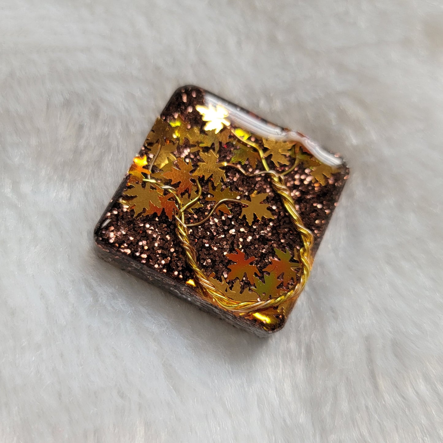 Square epoxy resin piece with fall trees scene made of gold colored jewelry wire and gold and holographic orange leaf shaped glitter.