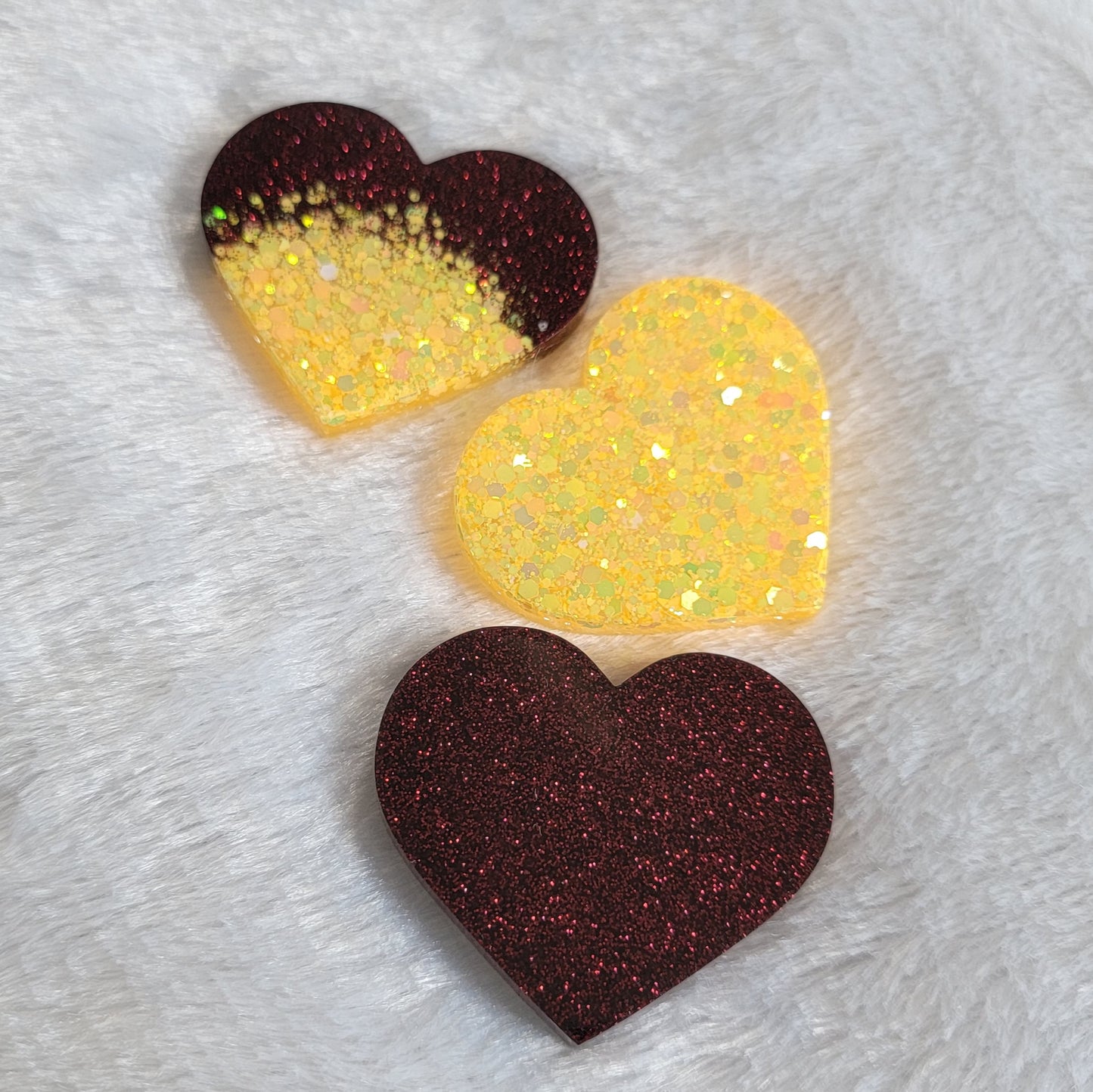 Three heart shaped epoxy resin pieces, one dark red glitter, one bright yellow glitter, and one a combination of the dark red and bright yellow glitters.