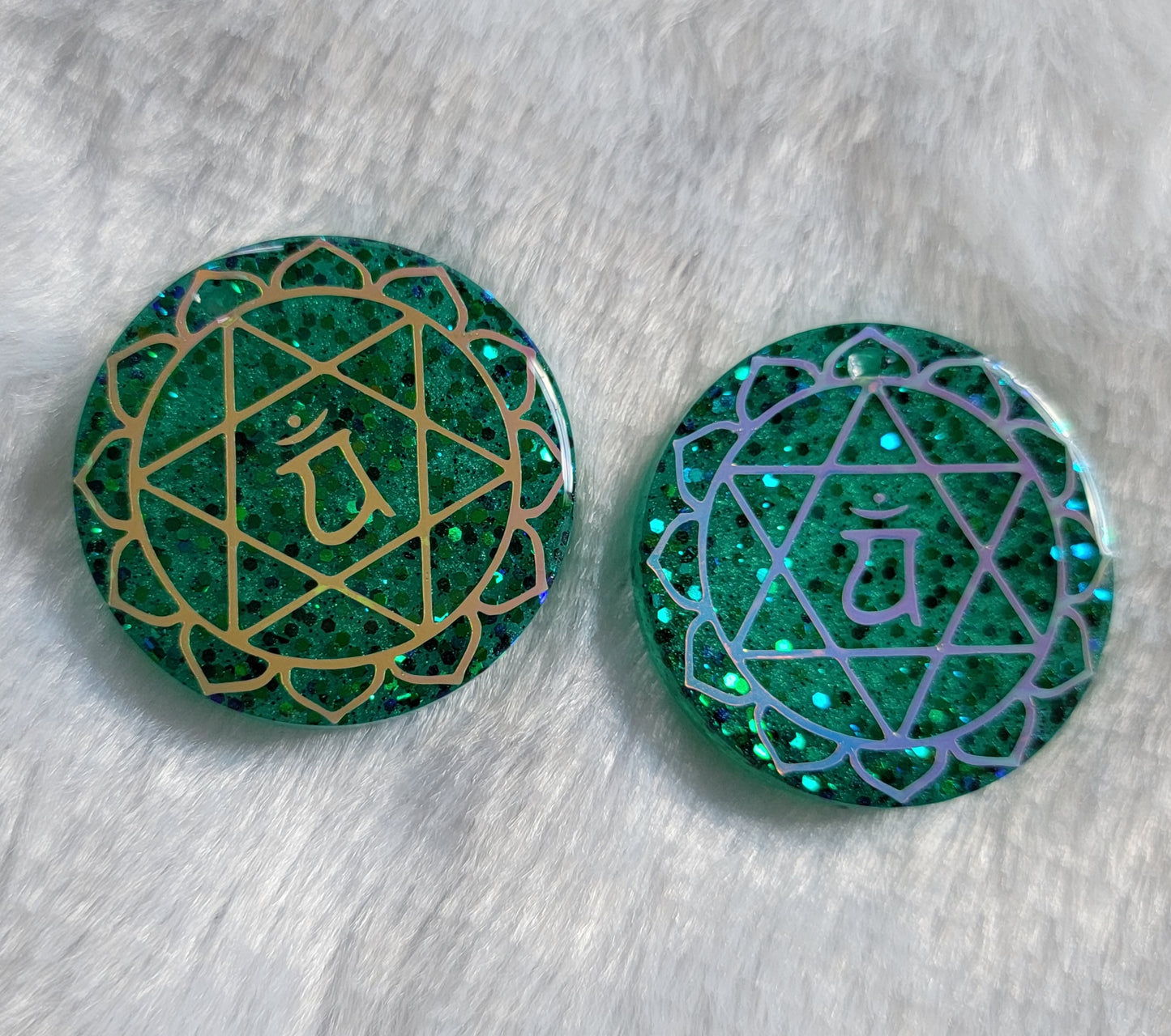 Two round epoxy resin pieces made with sparkling green glitter and pigment, each has heart chakra symbol made with vinyl - one golden one opal white.